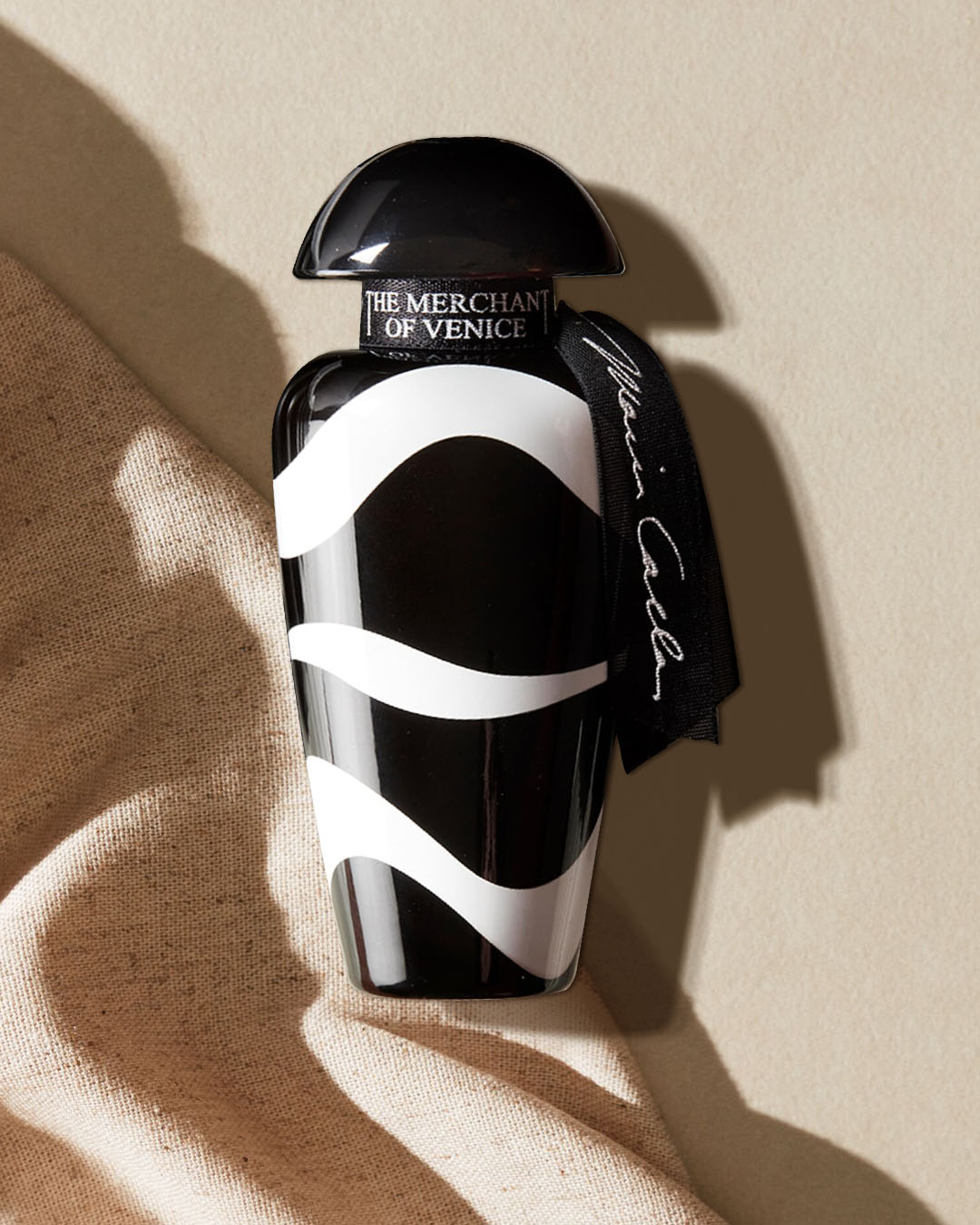 Flakon perfum The Merchant of Venice w czarno-białe paski, z czarną zakrętką i czarną wstążką z napisem "Maria Callas" na beżowym tle z tkaniny.