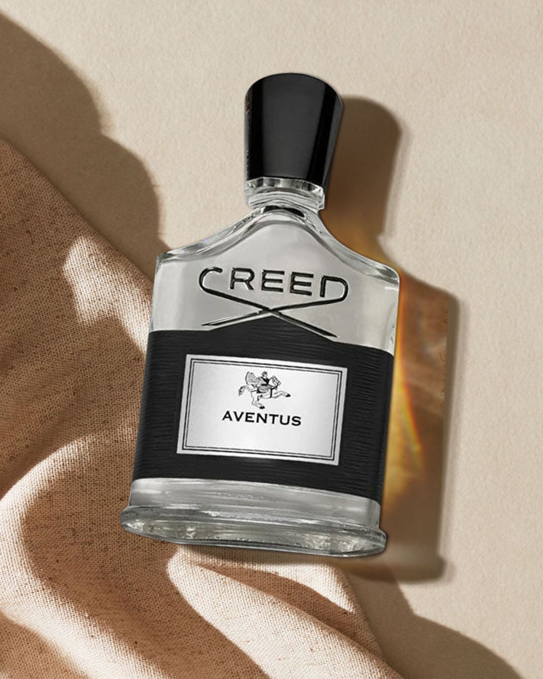 Flakon perfum Creed Aventus o pojemności 100 ml, z czarną etykietą i srebrnym napisem, leży na beżowej tkaninie, a obok niego widoczny jest cień.