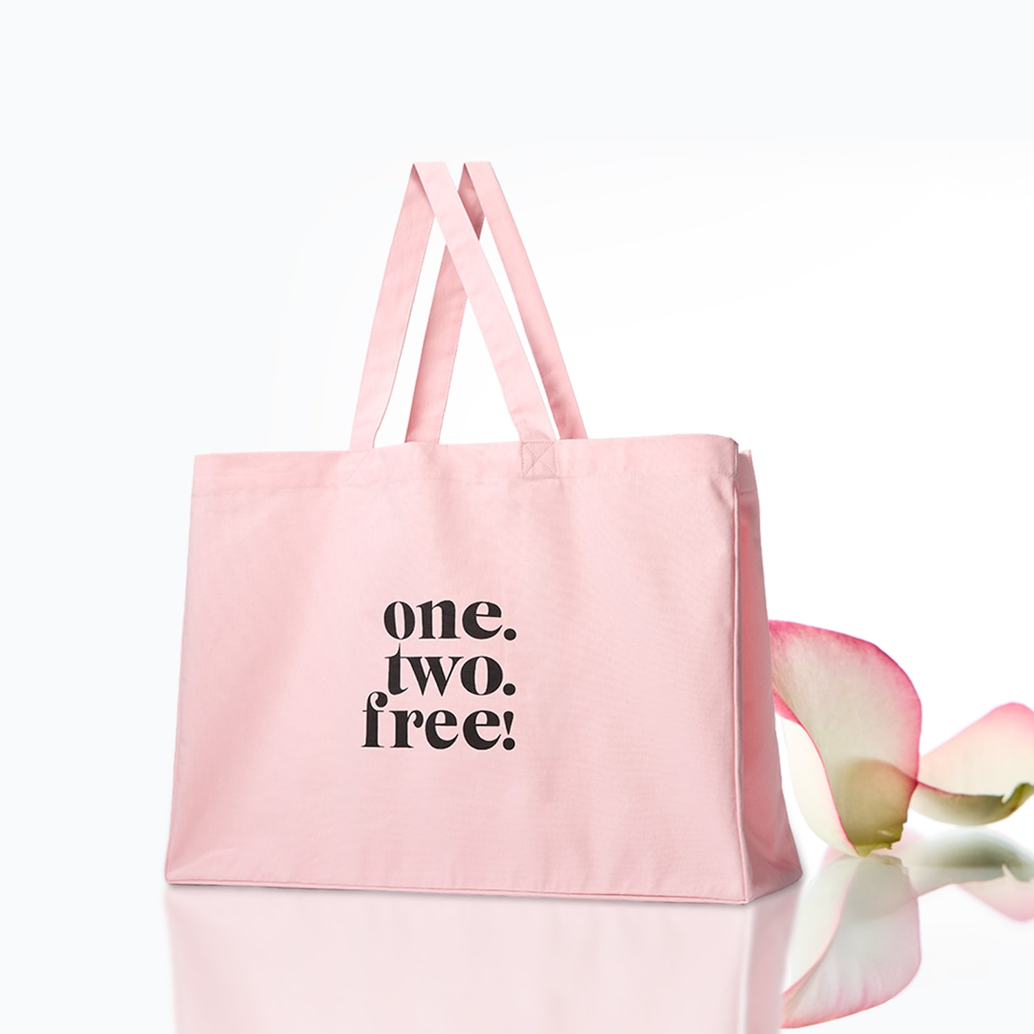 Eine hellrosa Stofftasche mit der Aufschrift „one. two. free!“ in schwarzer Schrift steht auf einer reflektierenden Oberfläche, daneben liegt ein rosafarbenes Blütenblatt.