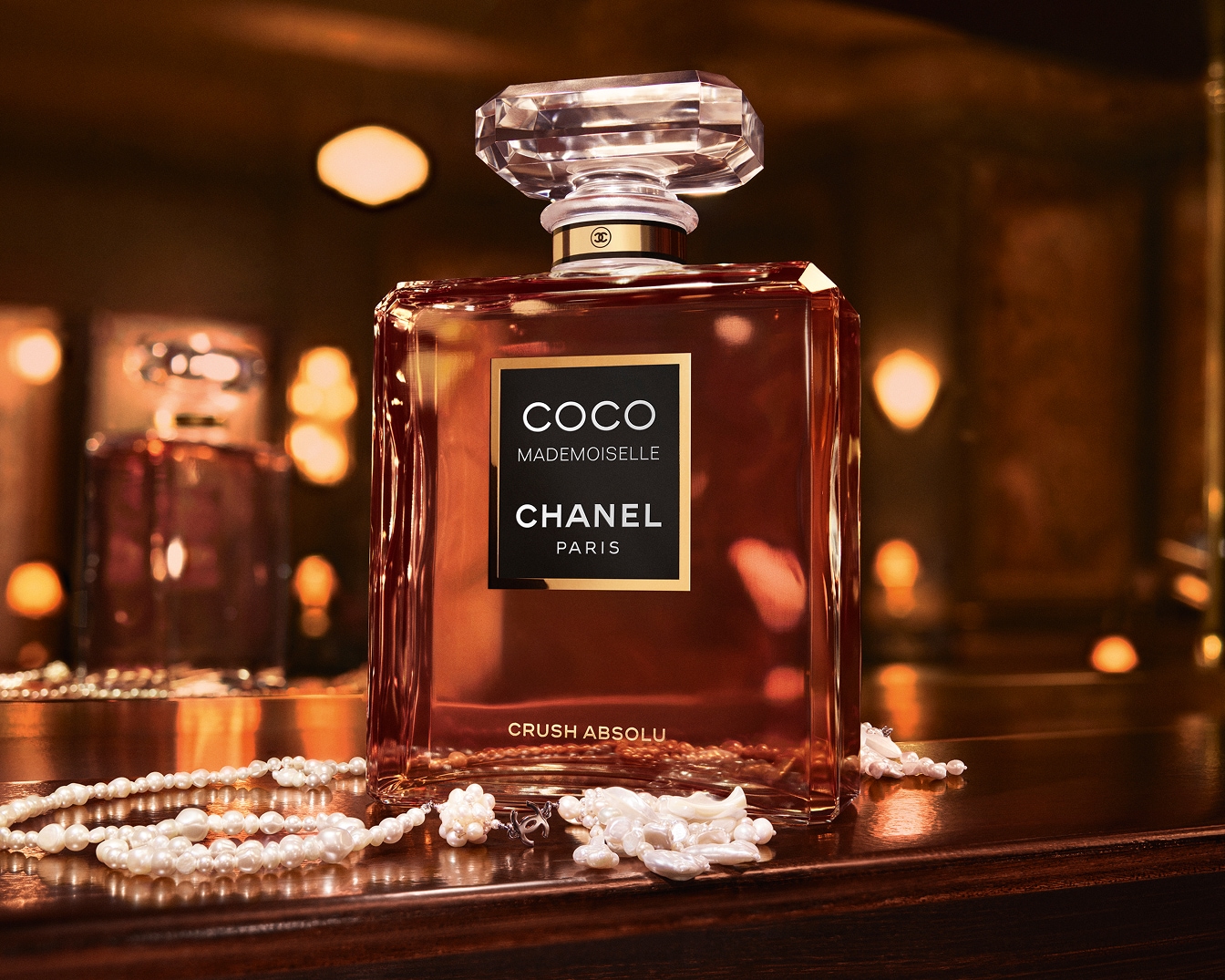 Un flacon elegant de parfum Coco Mademoiselle Chanel Paris, cu lichid de culoarea chihlimbarului, stă pe o suprafață lucioasă, înconjurat de șiraguri de perle albe, cu lumini calde estompate în fundal.