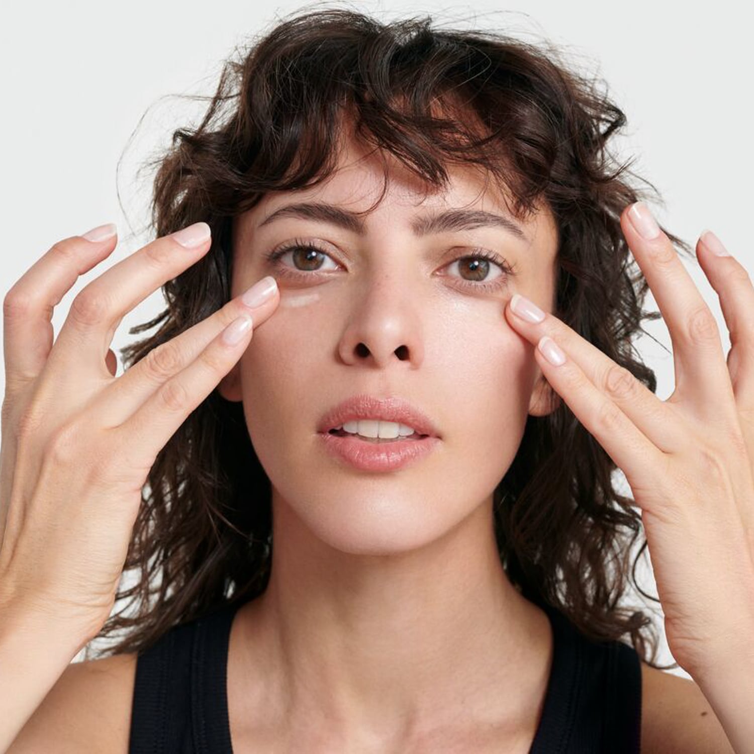 Eine Frau mit braunen, lockigen Haaren und einem schwarzen Oberteil trägt eine helle Creme unter ihren Augen auf, wobei sie ihre Finger sanft auf die Haut legt.