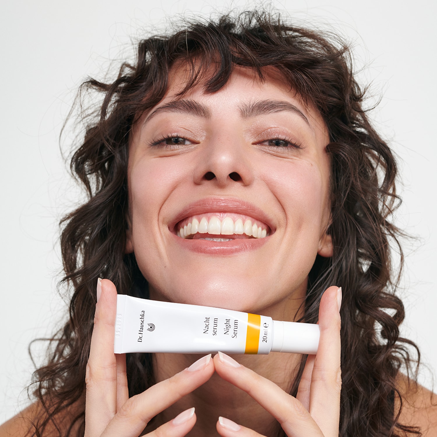 Eine lächelnde Frau mit lockigem, braunem Haar hält das Dr. Hauschka Nachtserum 20 ml in ihren Händen.
