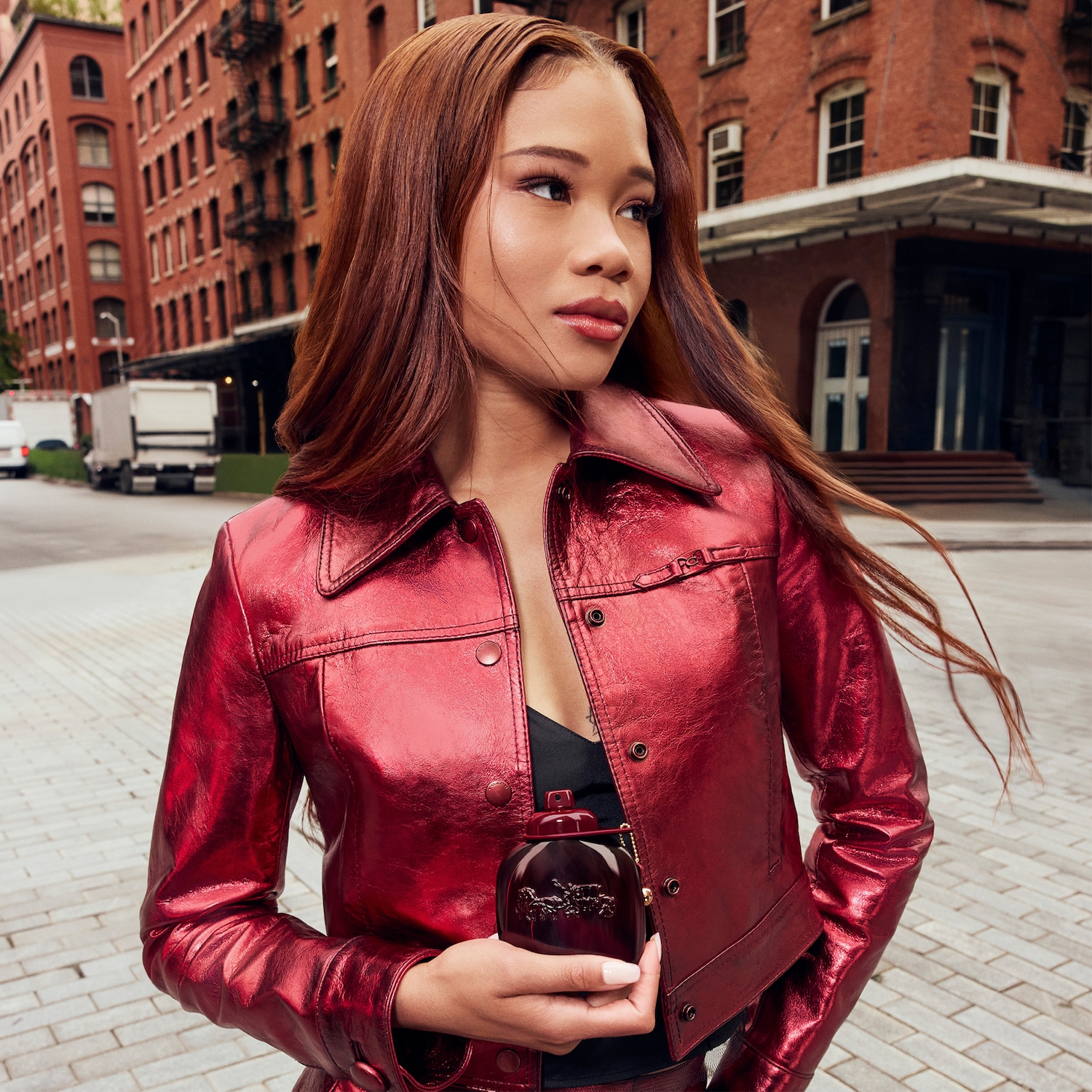 Une femme aux cheveux roux, vêtue d'une veste en cuir rouge vif, tient un flacon de parfum Coach rouge foncé avec un cheval en relief, sur fond de bâtiments en briques rouges.