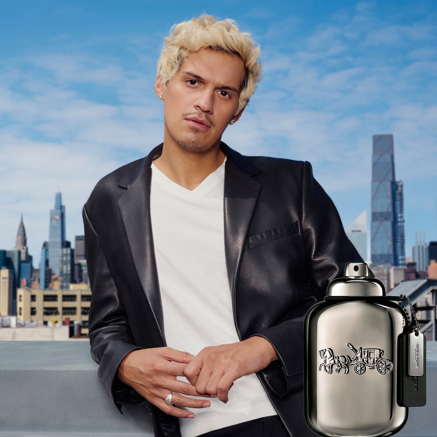 Un homme aux cheveux blonds décolorés, vêtu d'une veste en cuir noire et d'un t-shirt blanc, pose sur un toit avec la ville en arrière-plan, à côté d'une bouteille de parfum Coach for Men gris métallisé.