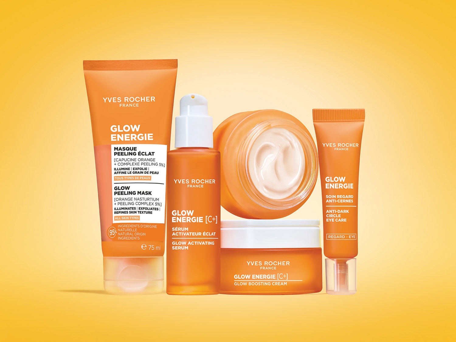 Zestaw Yves Rocher Glow Energie zawiera pomarańczową tubkę maski peelingującej, butelkę serum aktywującego blask, słoiczek kremu wzmacniającego blask oraz małą tubkę kremu pod oczy, wszystkie w pomarańczowych opakowaniach, ułożone na żółtym tle.