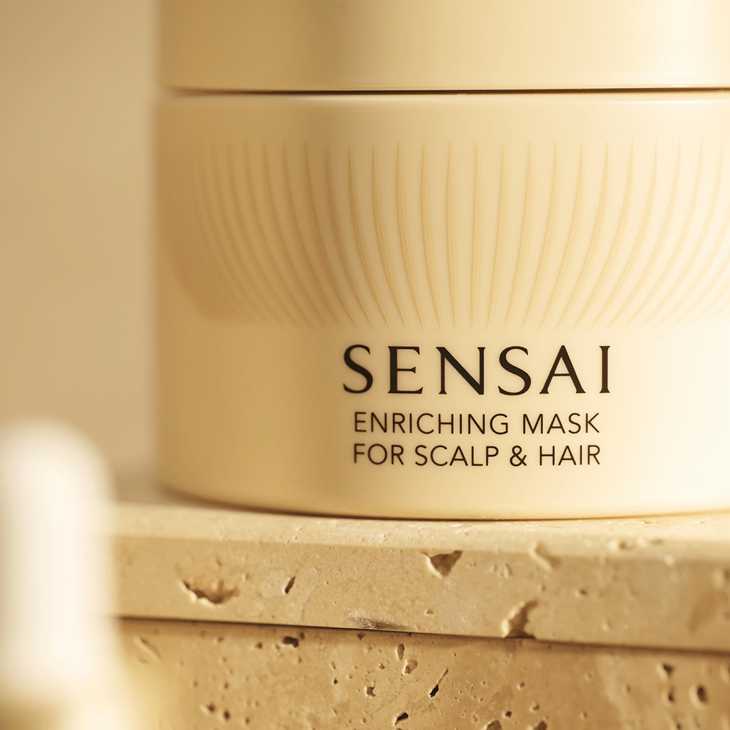 Le masque enrichissant Sensai pour le cuir chevelu et les cheveux est présenté dans un pot crème clair avec des rainures verticales, posé sur une surface texturée beige.