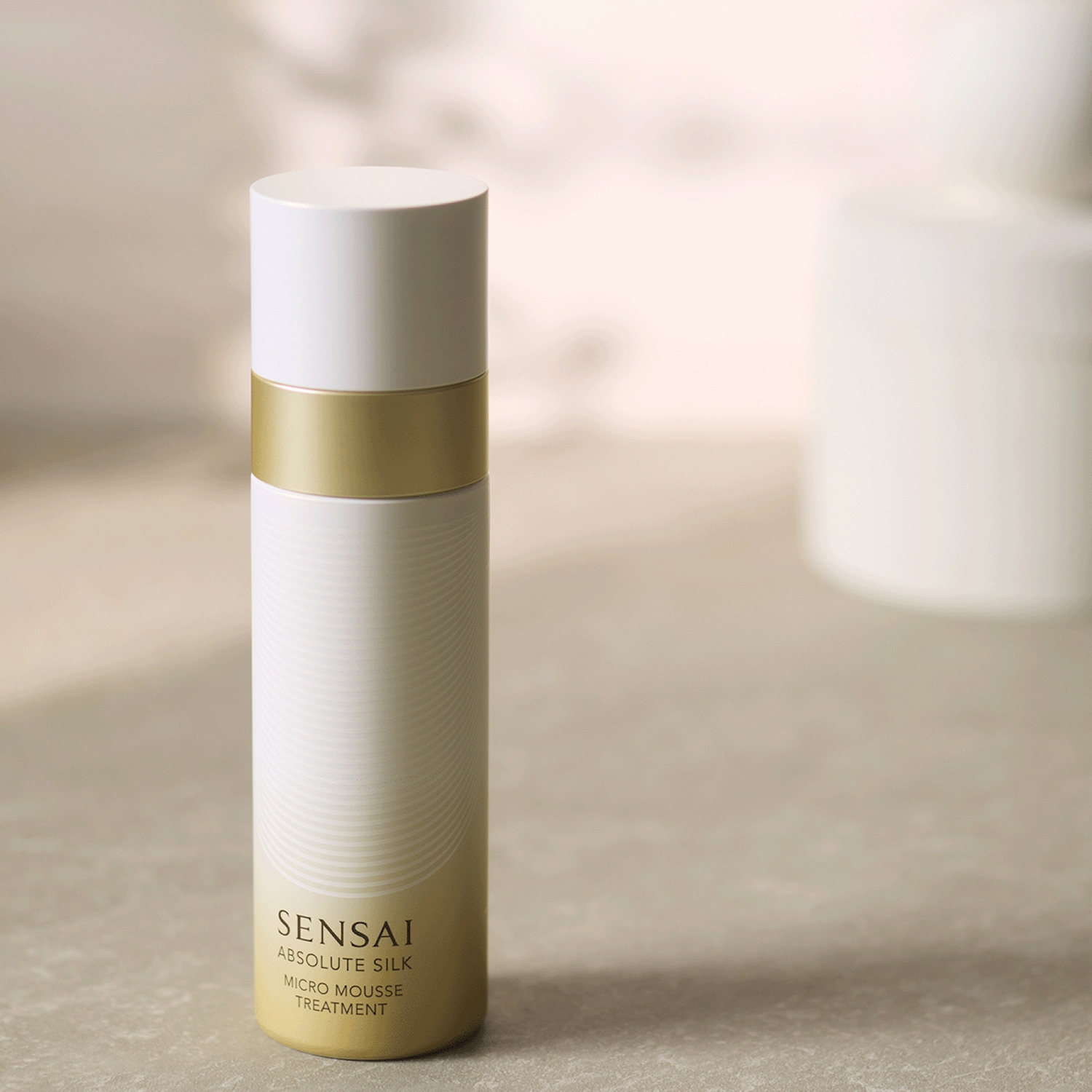 Le traitement micro-mousse Sensai Absolute Silk est présenté dans un flacon blanc élégant avec une bande dorée et un dégradé doré à la base, posé sur une surface grise texturée.