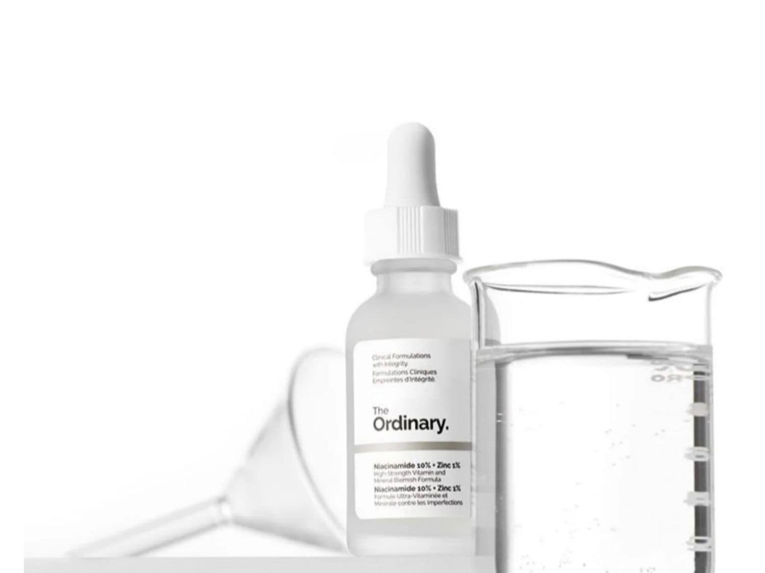 Bílá lahvička The Ordinary Niacinamide 10% + Zinc 1% s kapátkem stojí vedle skleněné kádinky s čirou tekutinou, což zdůrazňuje čistotu a vědecký přístup k péči o pleť.