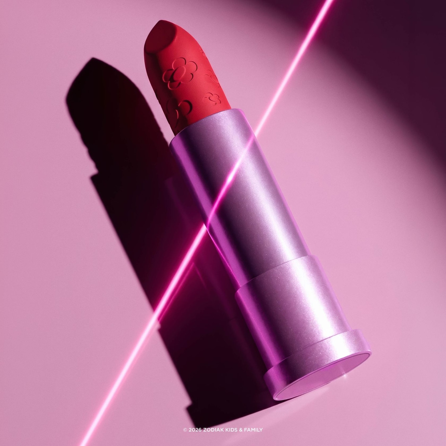 Un rouge à lèvres rouge vif avec des motifs de fleurs en relief est présenté dans un tube argenté, avec des reflets néon roses et une ombre portée sur un fond rose.