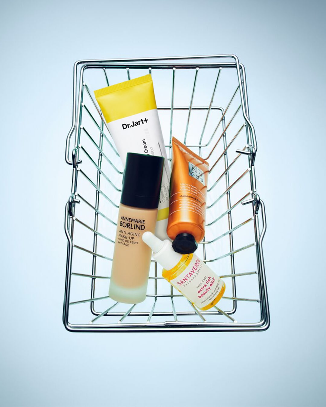 Een metalen winkelmandje gevuld met diverse huidverzorgings- en make-upproducten, waaronder Dr. Jart+ Cicapair Cream, Annemarie Börlind Anti-Aging Make-up, een oranje tube handcrème en Santaverde Extra Rich Beauty Elixir.