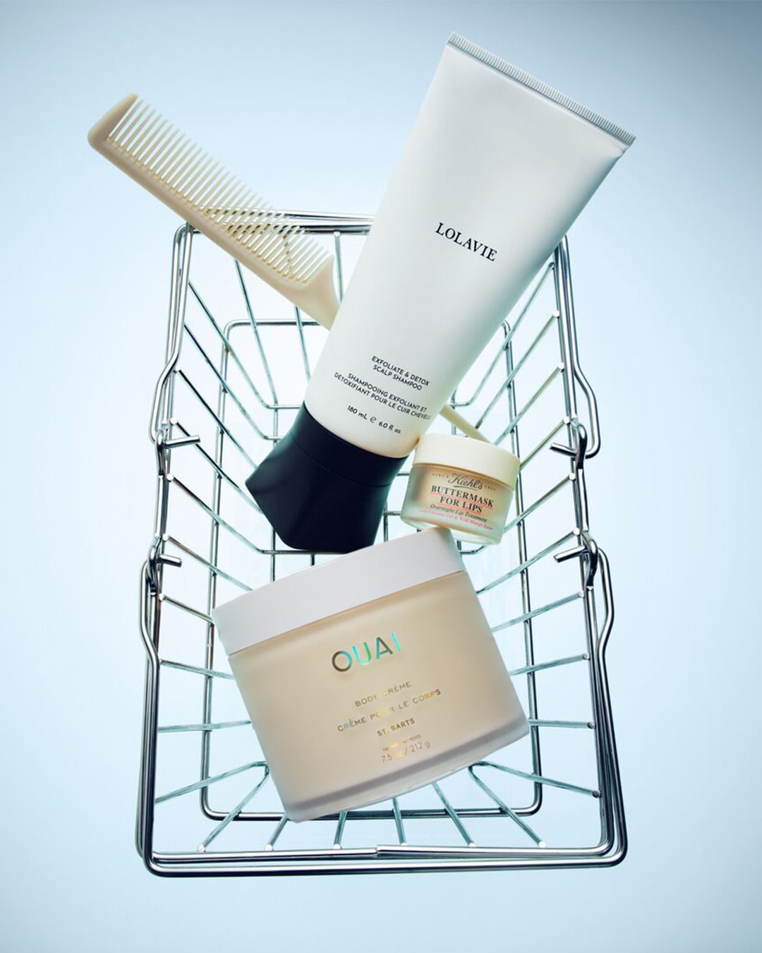 Een witte Lolavie Exfoliate & Detox Scalp Shampoo tube, een beige Ouai Body Crème pot en een kleine Kiehl's Buttermask for Lips pot liggen samen met een witte kam in een zilveren winkelmandje.