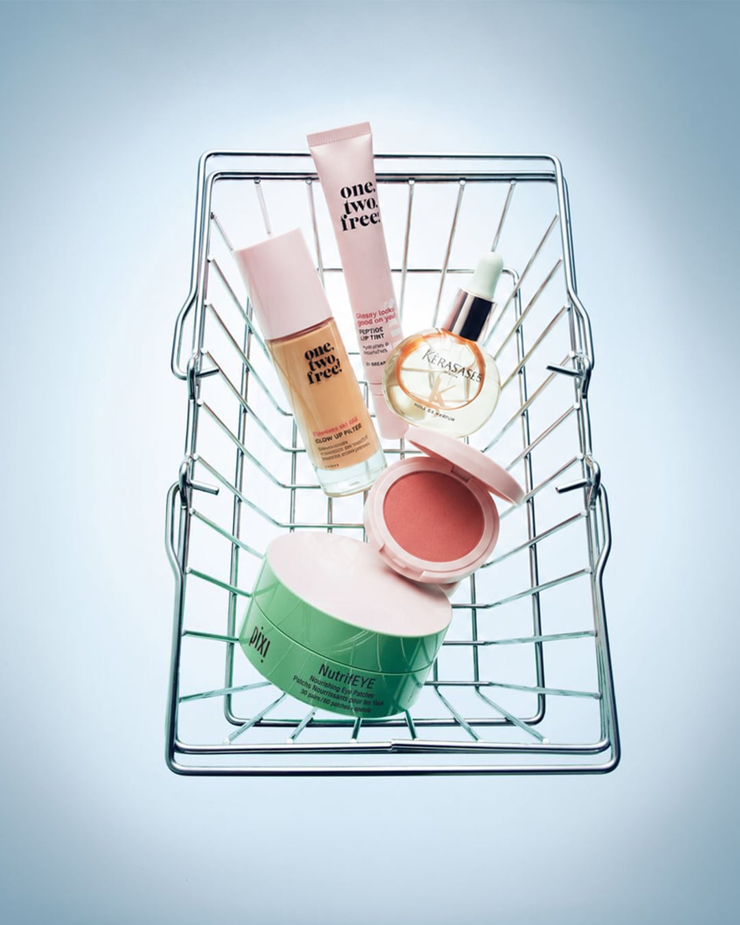 Een metalen winkelmandje met diverse make-up- en huidverzorgingsproducten, waaronder een foundation, liptint, oogpatches, blush en haarolie, alles in zachte pastelkleuren.