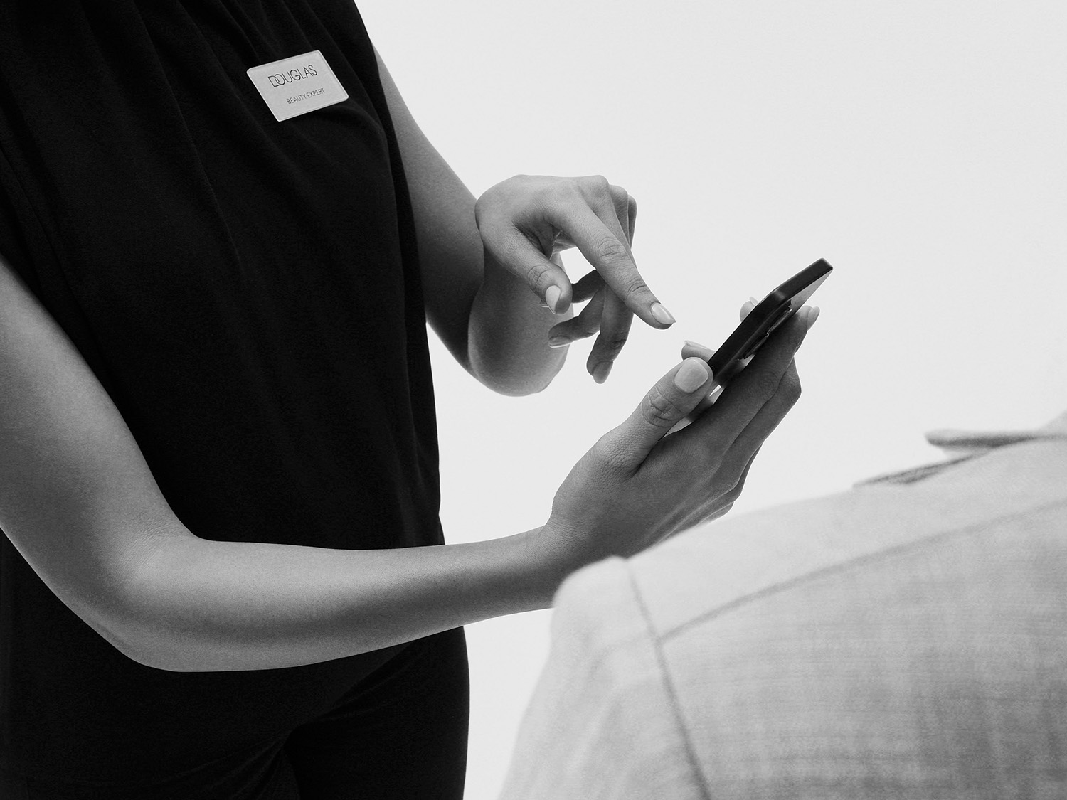 Douglas Beauty Expert v černé košili s jmenovkou Douglas Beauty Expert na hrudi, drží v rukou mobilní telefon a ukazuje na něj prstem, zatímco stojí vedle zákazníka.