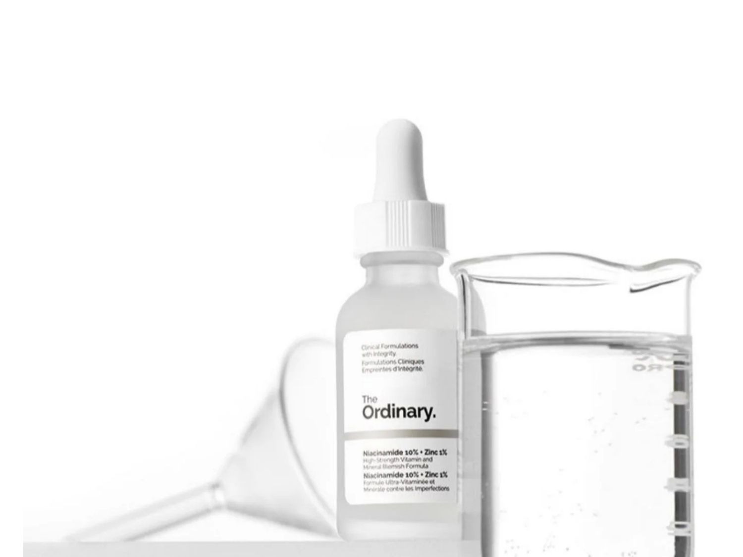 A The Ordinary Niacinamide 10% + Zinc 1% szérum egy fehér, cseppentős üvegben, mellette egy kémcsőben átlátszó folyadék, fehér háttér előtt.