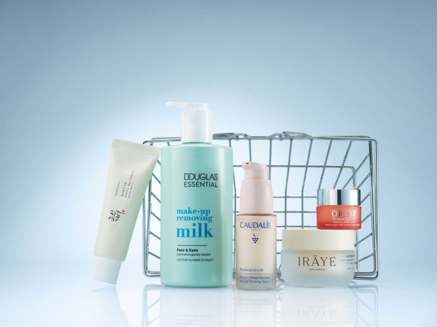 Pět kosmetických produktů je uspořádáno před drátěným košíkem, včetně bílé tuby krému na opalování Beauty of Joseon Relief Sun Rice Probiotics SPF50+ PA++++, světle modré lahvičky odličovacího mléka Douglas Essential s pumpičkou, béžového séra Caudalie Resveratrol-Lift Instant Firming Serum s pumpičkou, oranžového krému Clinique All About Eyes Rich a bílého krému Iräye The Rich Cream.