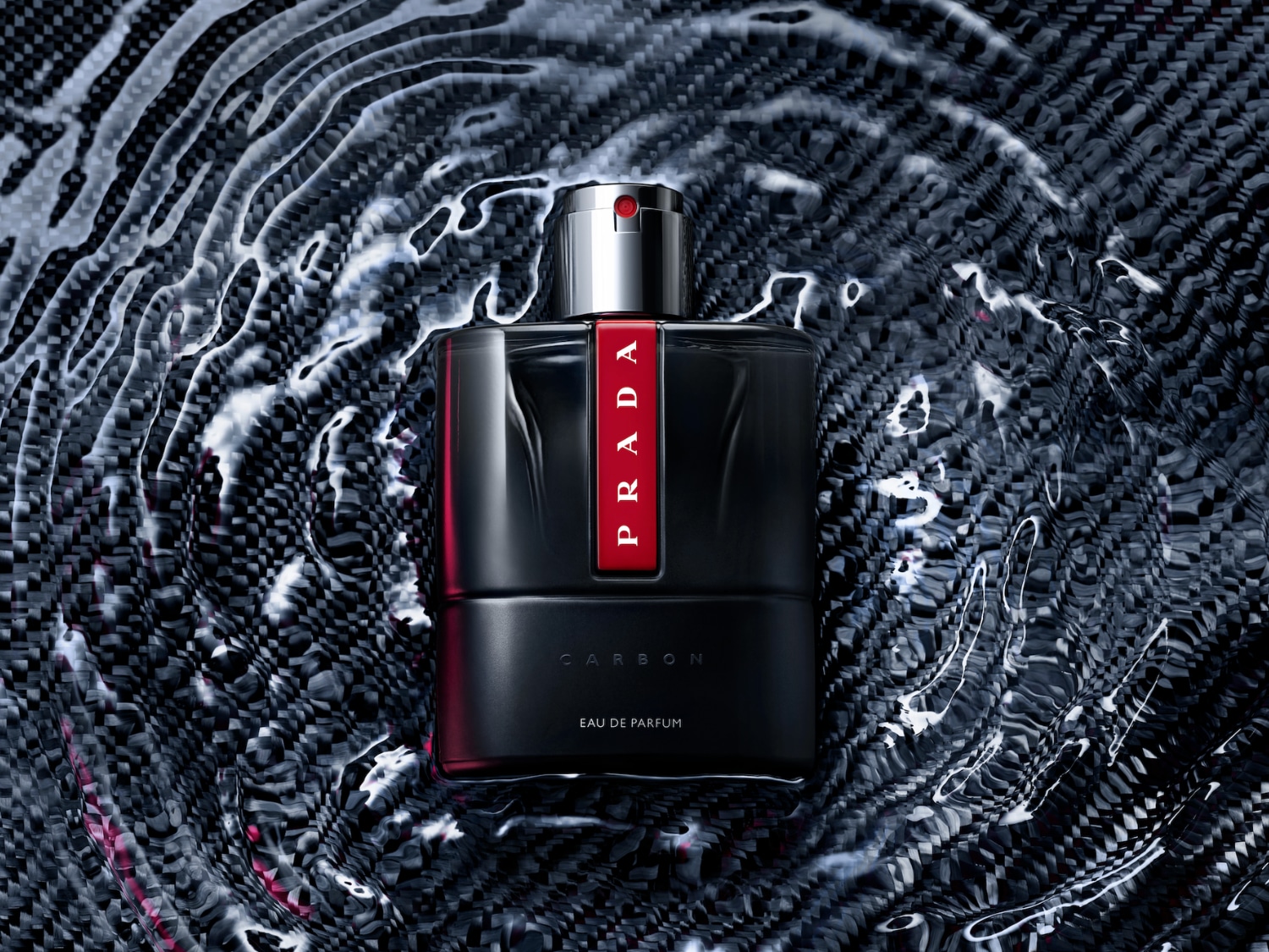 Prada Carbon Eau de Parfum pro muže v elegantním černém flakonu s červeným pruhem a stříbrným uzávěrem, obklopený vlnící se vodou na tmavém pozadí.