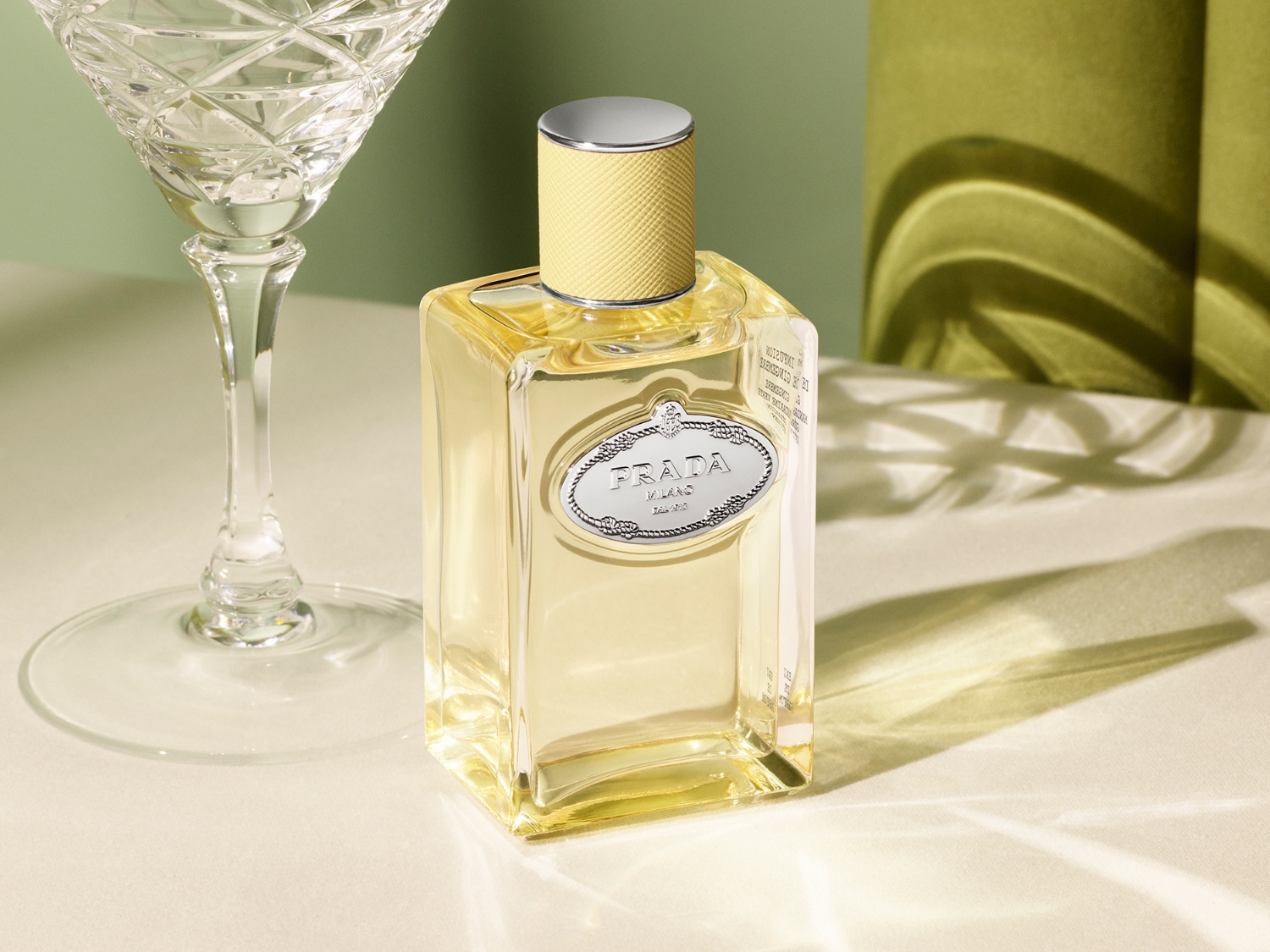 Prada Infusion d'Iris Eau de Parfum je elegantní vůně v hranaté skleněné lahvičce se stříbrným uzávěrem a žlutým texturovaným hrdlem, vedle níž stojí broušená sklenice na víno, vše na světle zeleném pozadí.