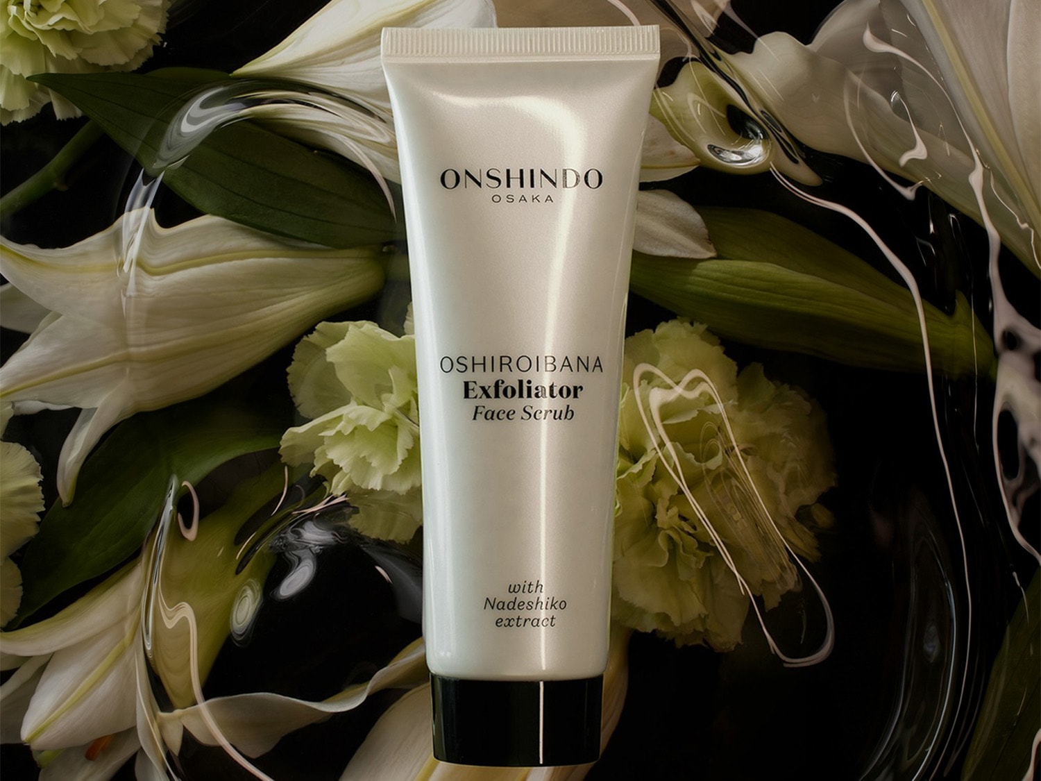 Biała tuba Onshindo Osaka Oshiroibana Exfoliator Face Scrub z czarną nakrętką, zawierająca ekstrakt z Nadeshiko, leży na ciemnym tle z białymi kwiatami lilii i zielonymi liśćmi, otoczona przezroczystymi, falującymi smugami.