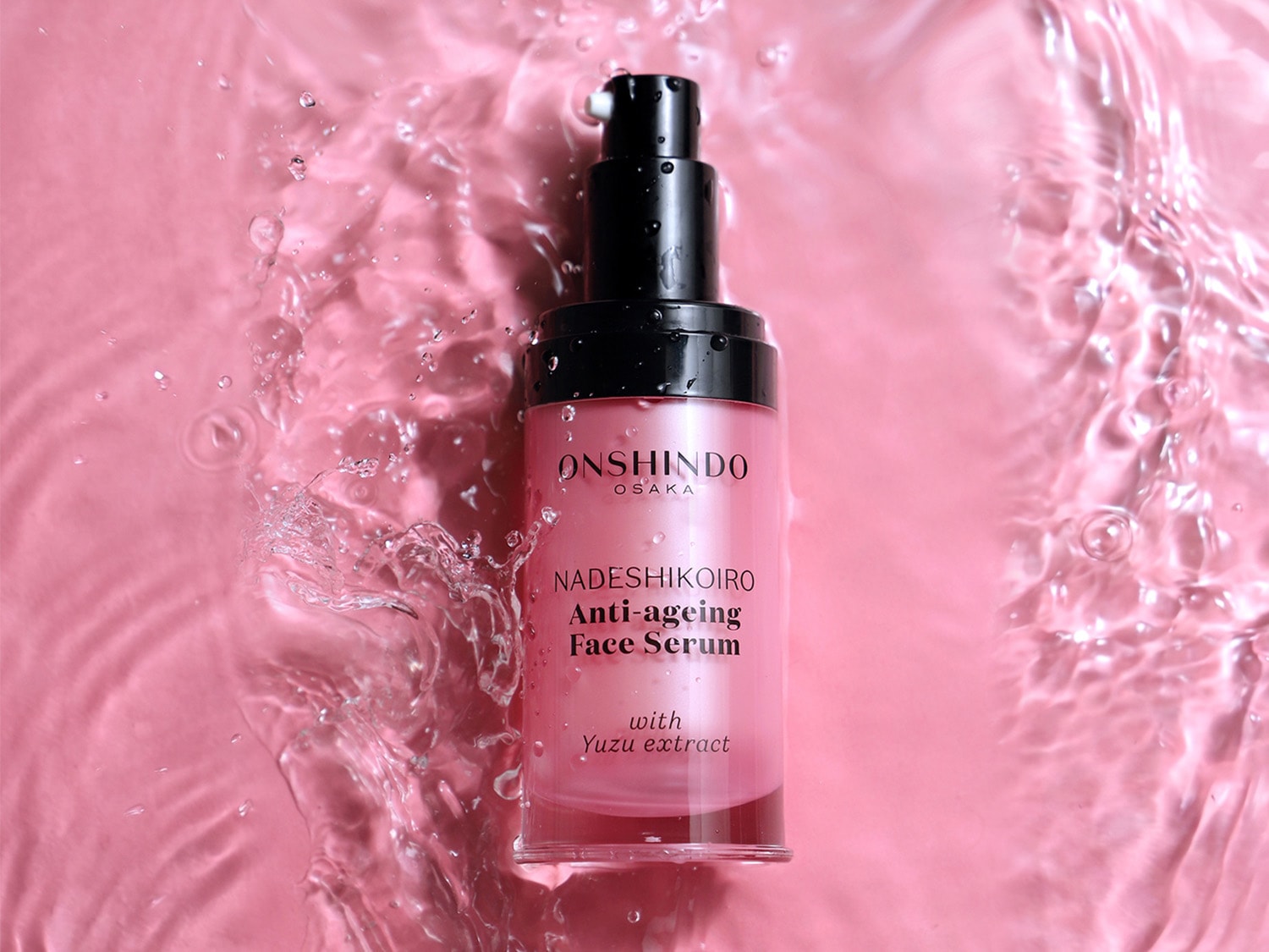 Serum do twarzy Onshindo Osaka Nadeshikoiro Anti-ageing z ekstraktem z yuzu w różowej butelce z czarną pompką, otoczone wodą, która rozpryskuje się na różowym tle.