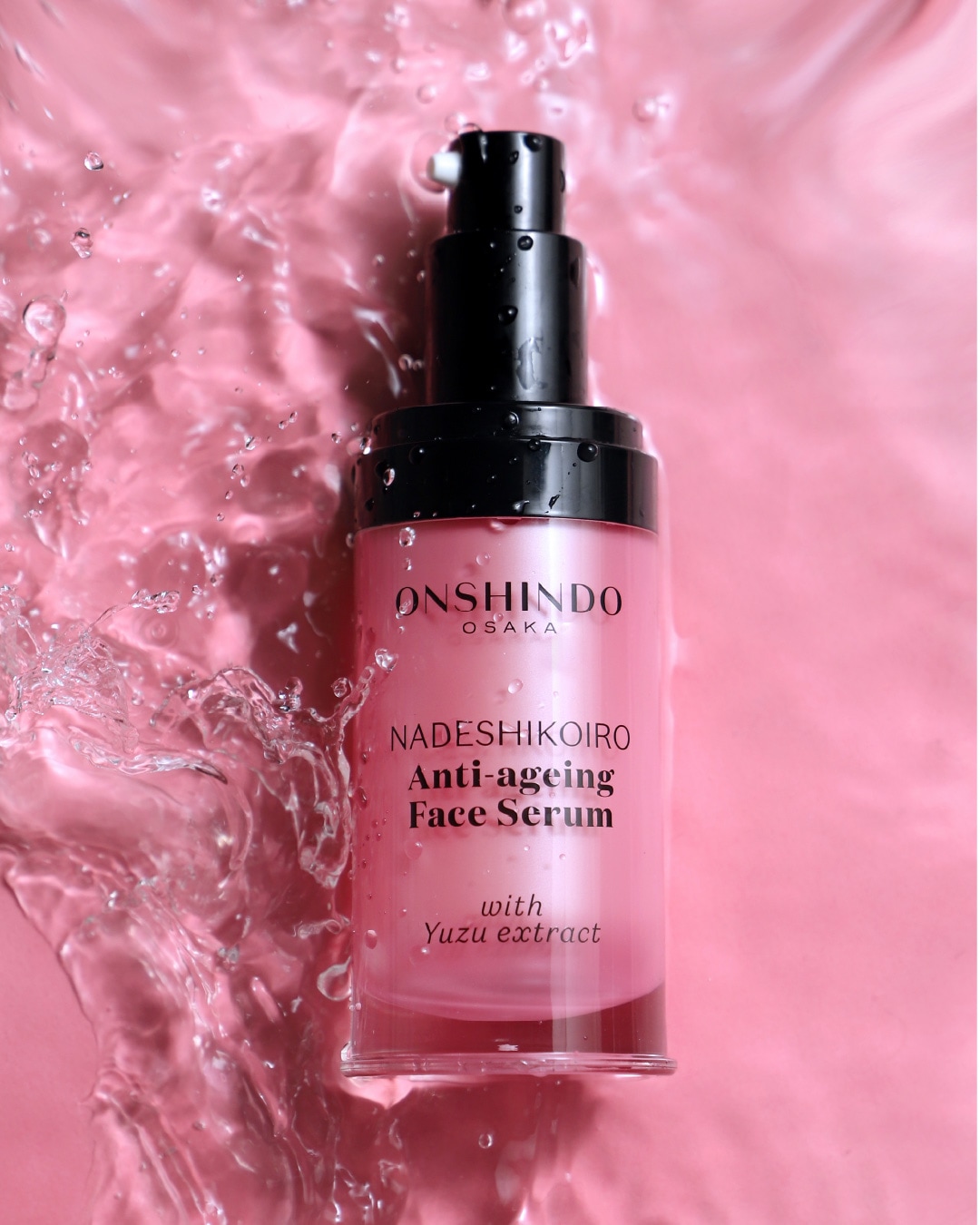 Serum do twarzy Onshindo Osaka Nadeshikoiro Anti-ageing z ekstraktem z Yuzu w różowej butelce z czarną pompką, otoczone rozbryzgami wody na różowym tle.
