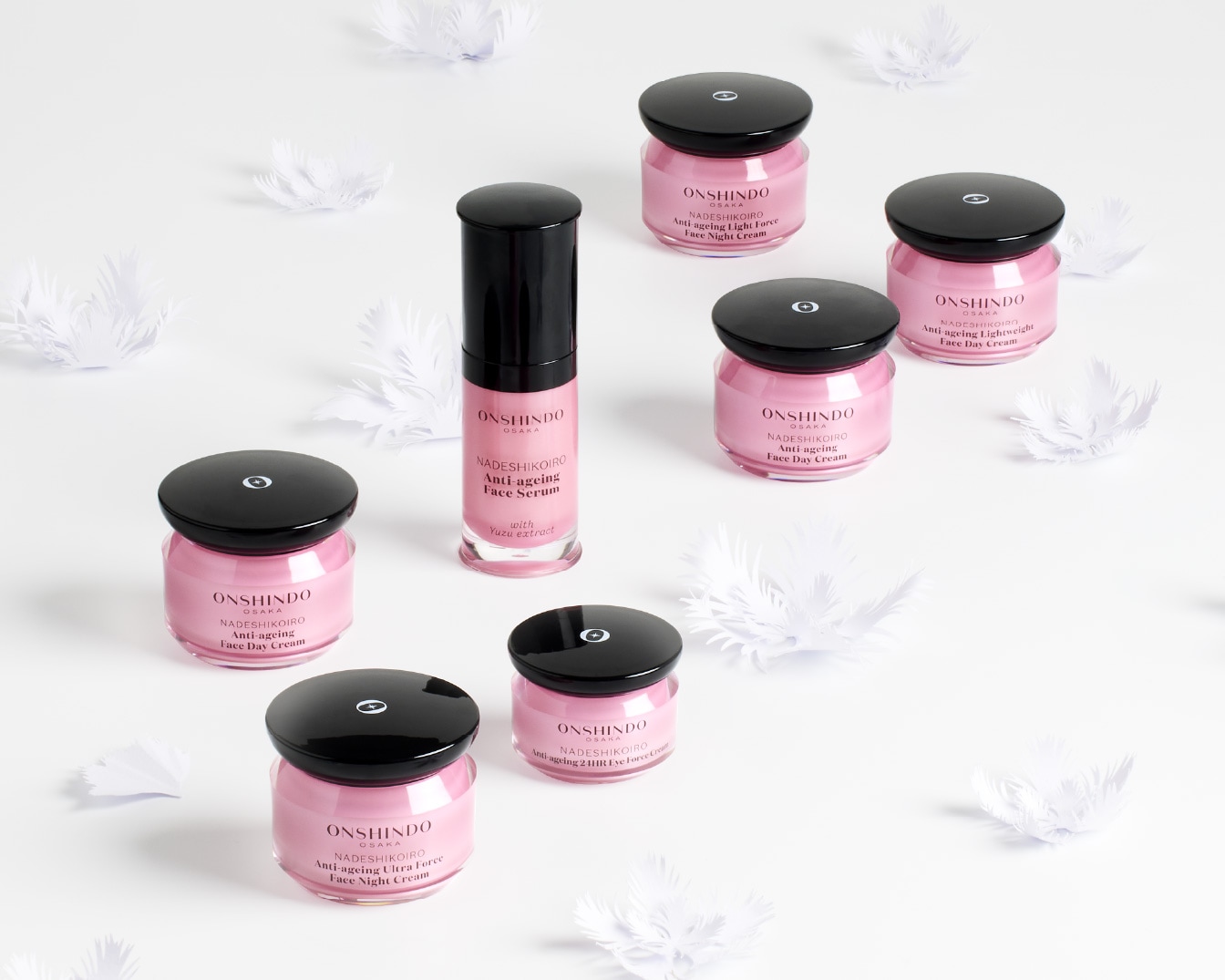 Odkryj kolekcję Onshindo Osaka Nadeshikoiro, w tym serum do twarzy z ekstraktem z yuzu, kremy na dzień i na noc, oraz krem pod oczy, wszystkie w eleganckich różowych słoiczkach z czarnymi zakrętkami, otoczone białymi piórkami.