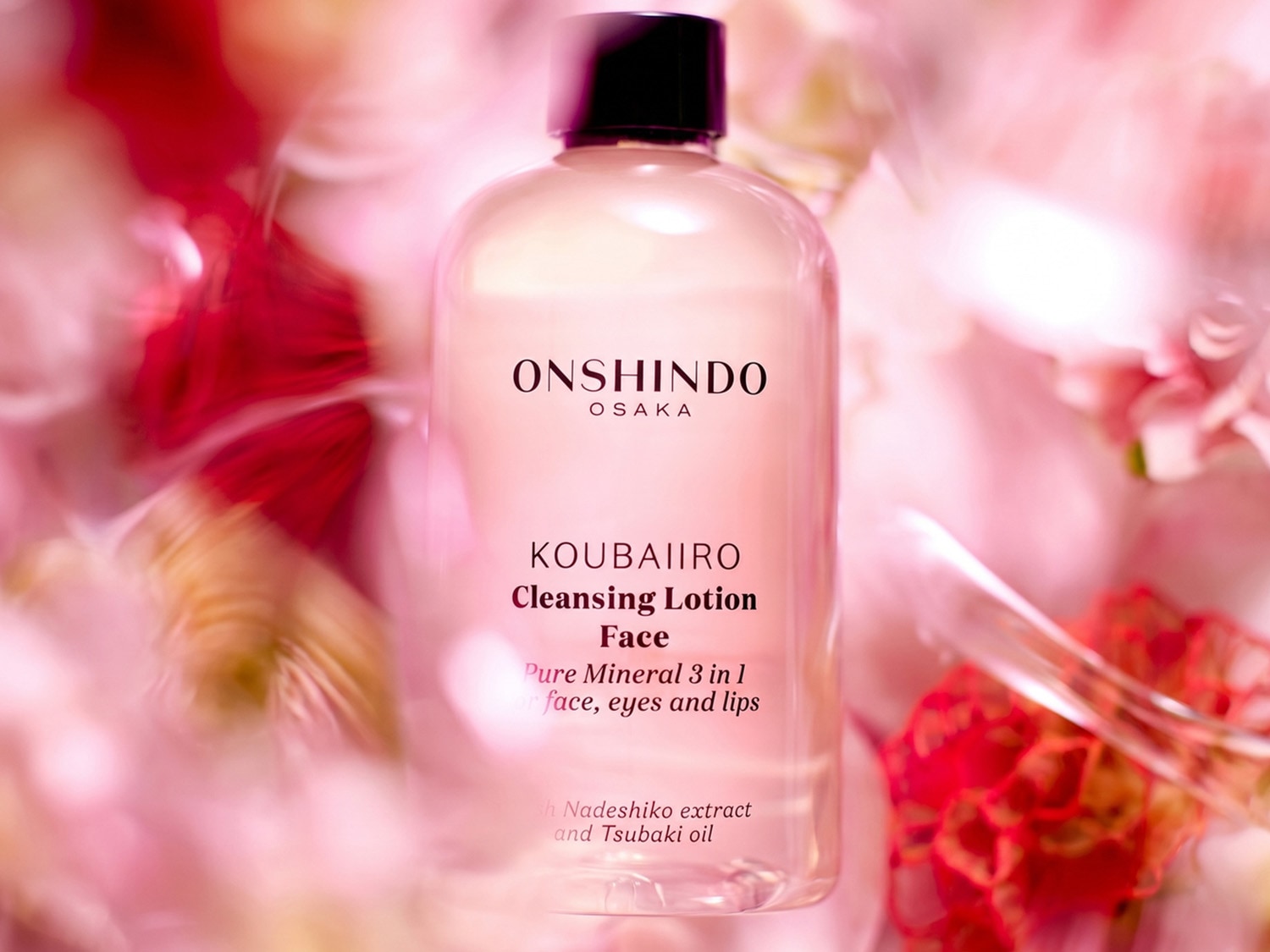 Przezroczysta butelka ONSHINDO KOUBAIIRO Cleansing Lotion Face z czarną nakrętką, otoczona rozmytymi różowymi i czerwonymi płatkami, zawiera płyn do demakijażu 3 w 1 do twarzy, oczu i ust z ekstraktem z Nadeshiko i olejkiem Tsubaki.
