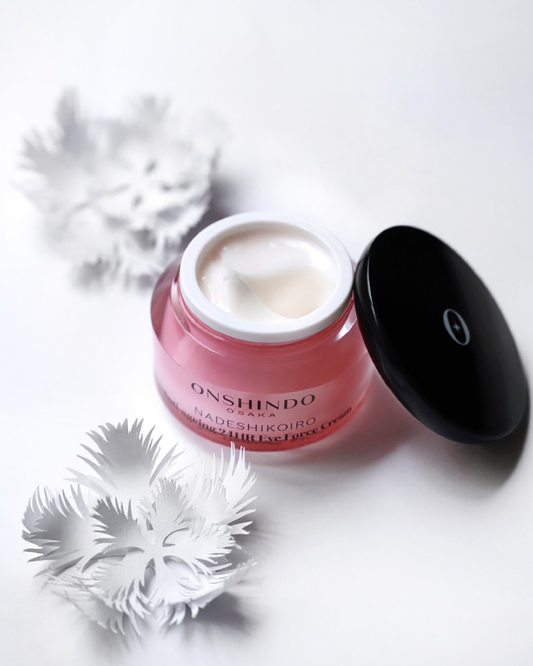 Odkryj krem pod oczy Onshindo Osaka Nadeshikoiro Anti-Ageing 24HR Eye Force Cream w różowym słoiczku z czarną zakrętką, otoczony białymi, papierowymi kwiatami.