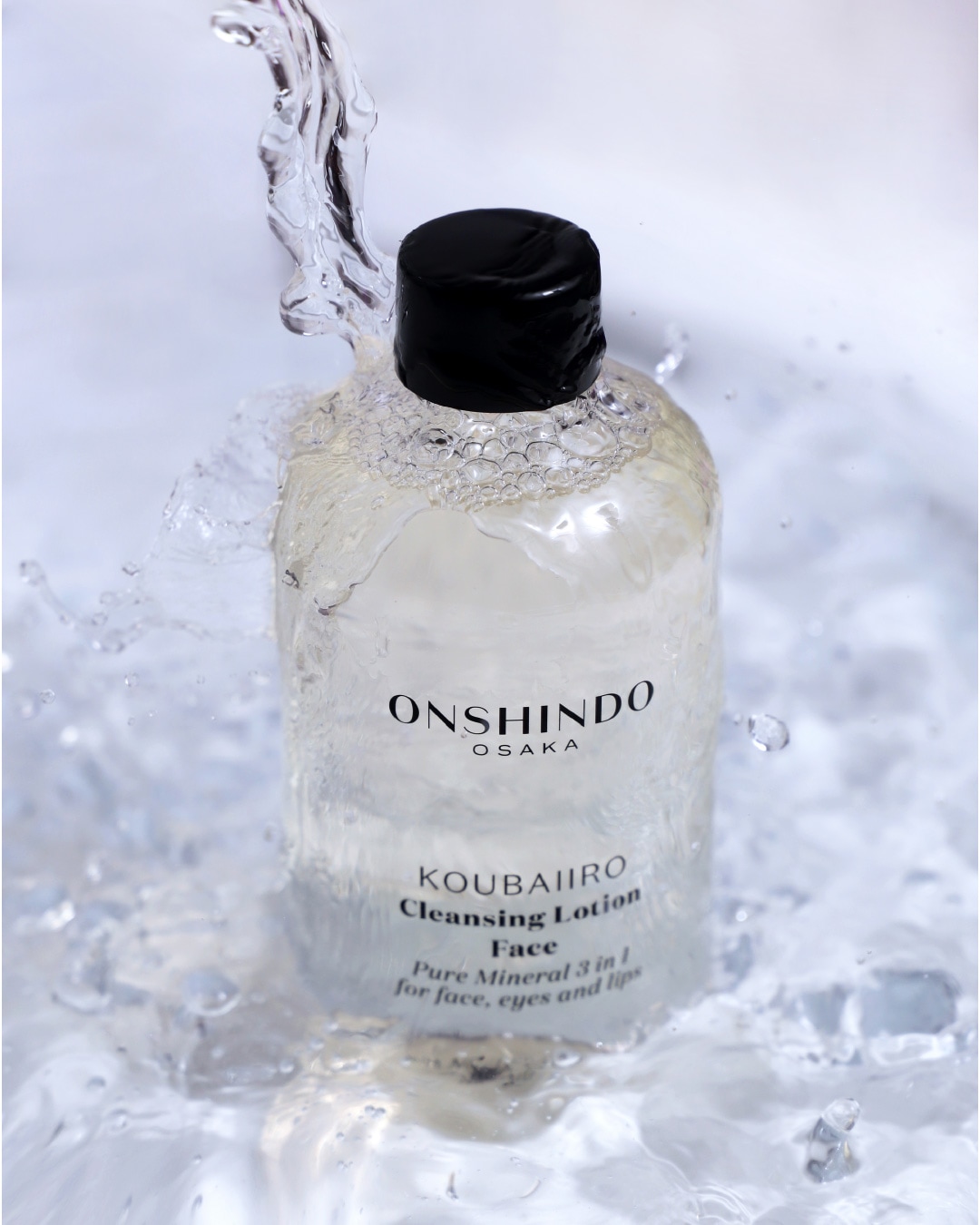 Przezroczysta butelka ONSHINDO OSAKA KOUBAIIRO Cleansing Lotion do twarzy, oczu i ust, z czarną zakrętką, jest otoczona rozbryzgami wody, co podkreśla jej odświeżające właściwości.