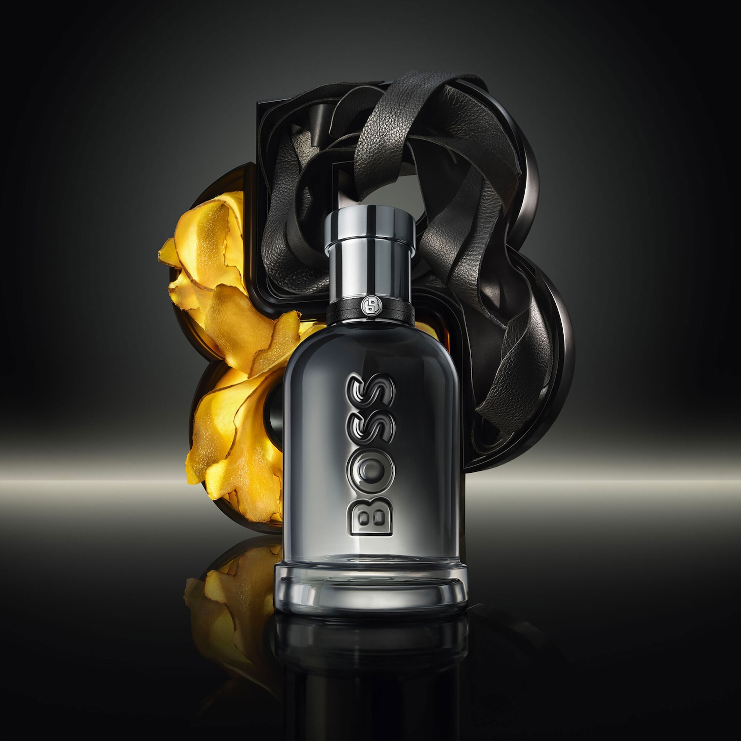 Fľaša parfému Boss The Scent Le Parfum pre mužov je zobrazená v elegantnej čiernej farbe s lesklým strieborným uzáverom a nápisom BOSS, doplnená o kožený remienok a zlaté lupienky v pozadí.