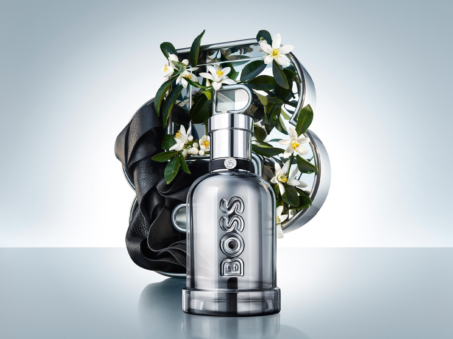 Strieborná fľaštička BOSS The Scent Le Parfum pre neho, s čiernym koženým detailom a bielymi kvetmi pomarančovníka, je umiestnená na lesklom povrchu.