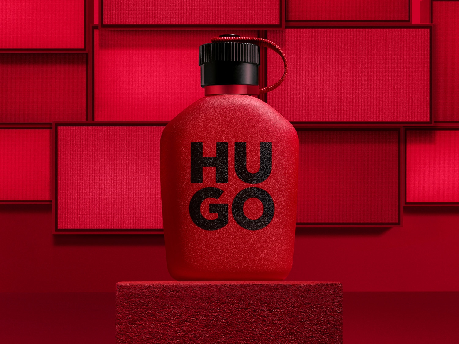 Červená fľaša parfému Hugo Boss s čiernym uzáverom a nápisom "HUGO" v čiernej farbe, umiestnená na červenom podstavci s červeným pozadím.