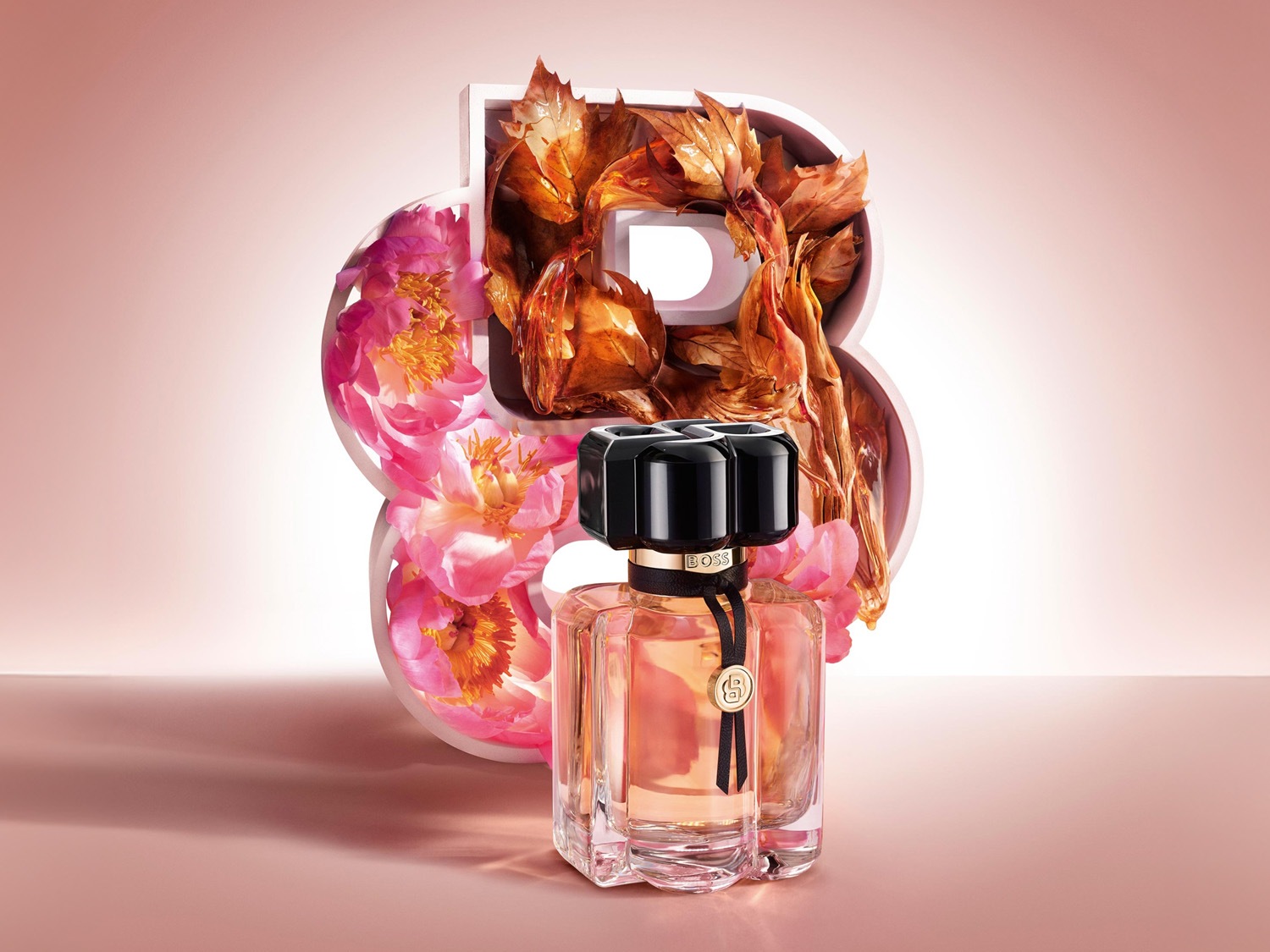 Parfumovaná voda Boss The Scent Le Parfum pre ženy v hranatom flakóne s čiernym uzáverom a stuhou, s ružovými kvetmi a jantárovými listami v pozadí.