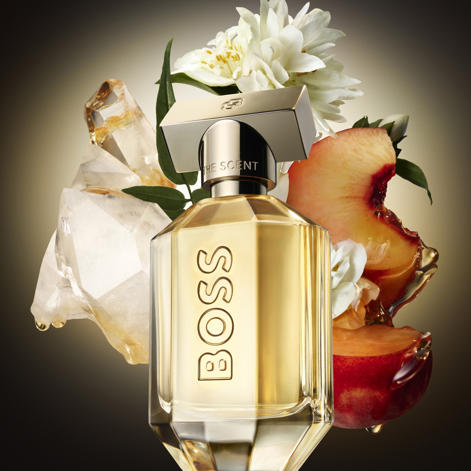 Parfumovaná voda Boss The Scent pre ženy v elegantnom flakóne so zlatým uzáverom, obklopená bielymi kvetmi, broskyňou a kryštálmi, vytvára zmyselnú a lákavú atmosféru.