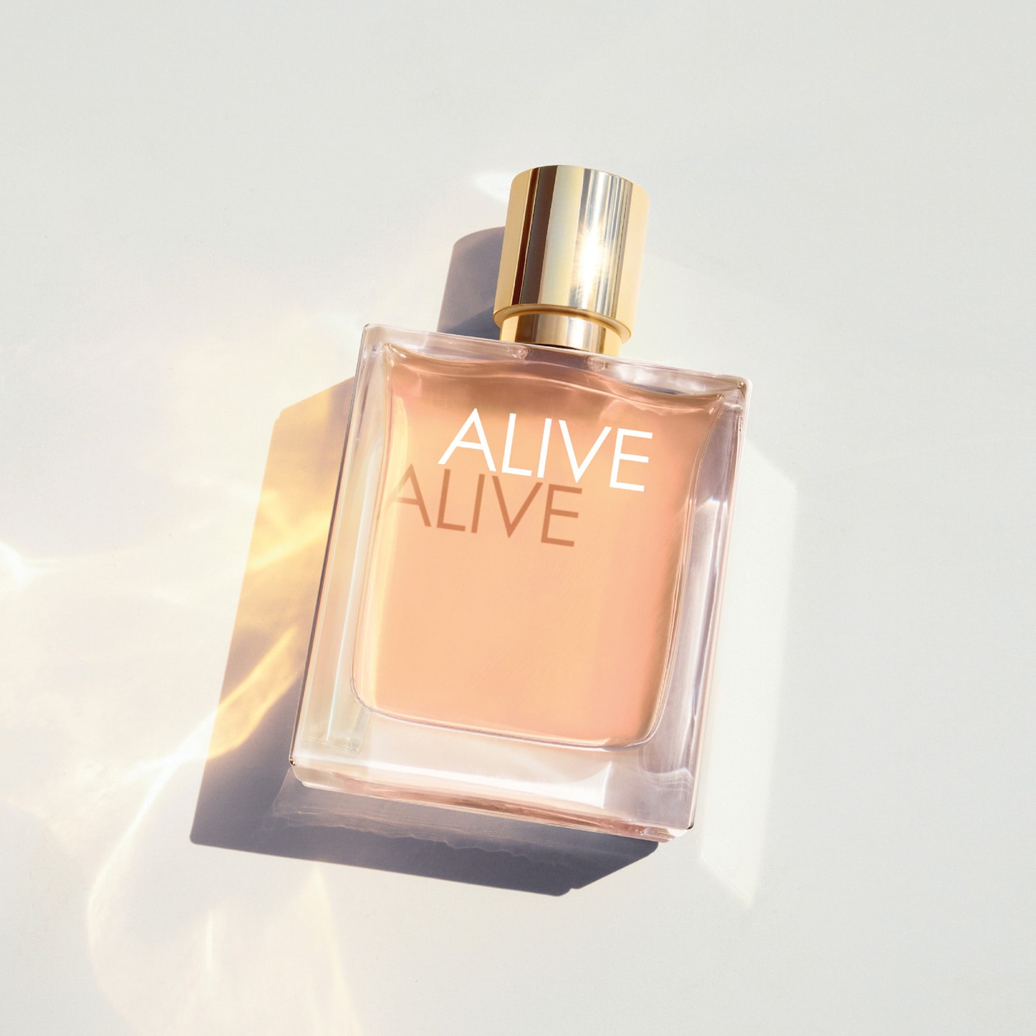 Parfum Alive v obdĺžnikovom flakóne s broskyňovou tekutinou a zlatým uzáverom, s nápisom ALIVE v bielom texte, leží na bielom povrchu s tieňmi a odleskami.