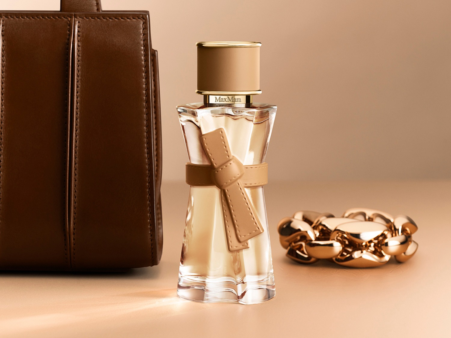 Un frasco de perfume Max Mara de color ámbar claro con un lazo de cuero beige, junto a un bolso de cuero marrón y una pulsera dorada, sobre un fondo beige.