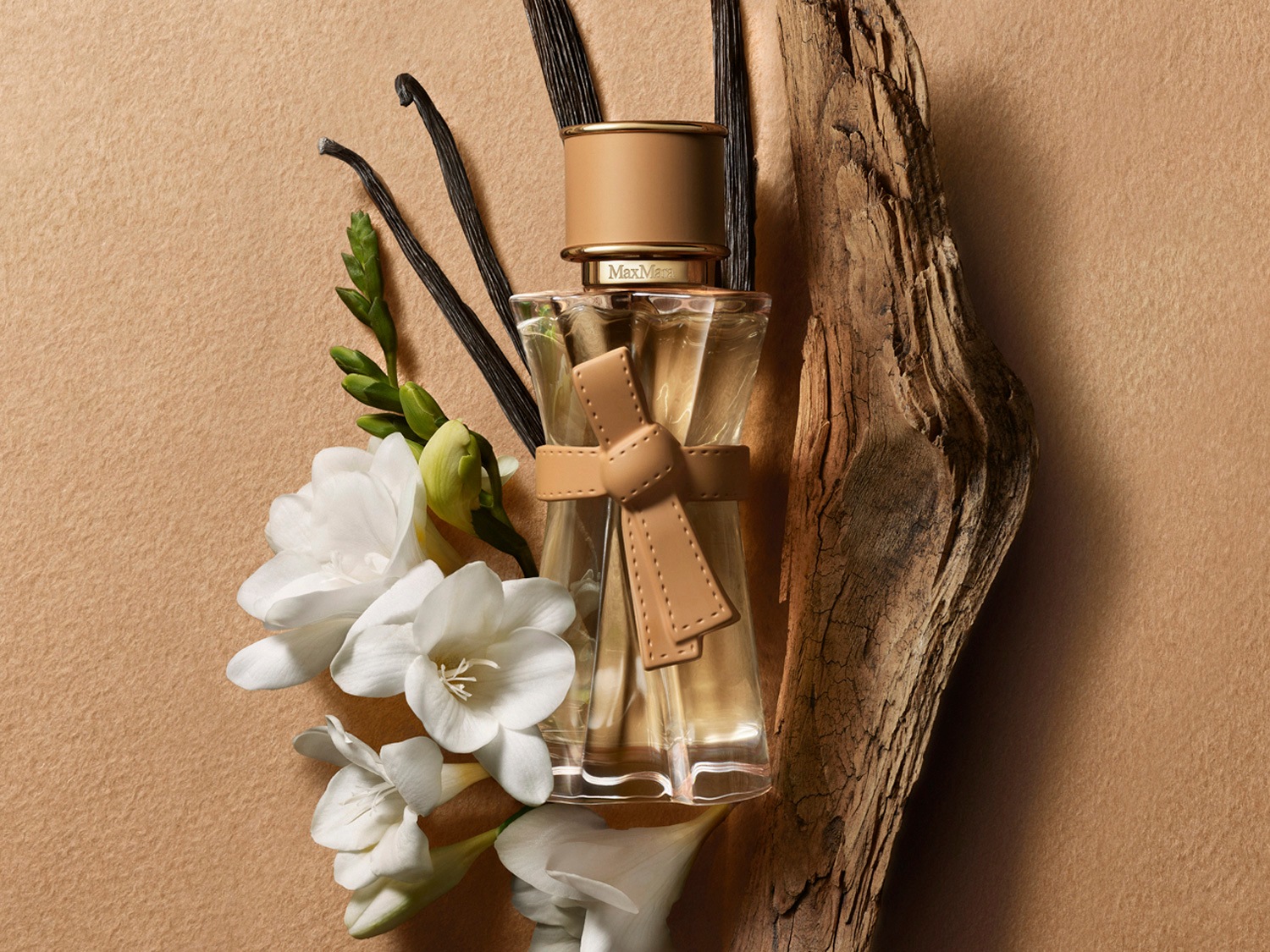 Un frasco de perfume Max Mara con un lazo de cuero beige, rodeado de vainas de vainilla, flores blancas de fresia y un trozo de madera, sobre un fondo beige.