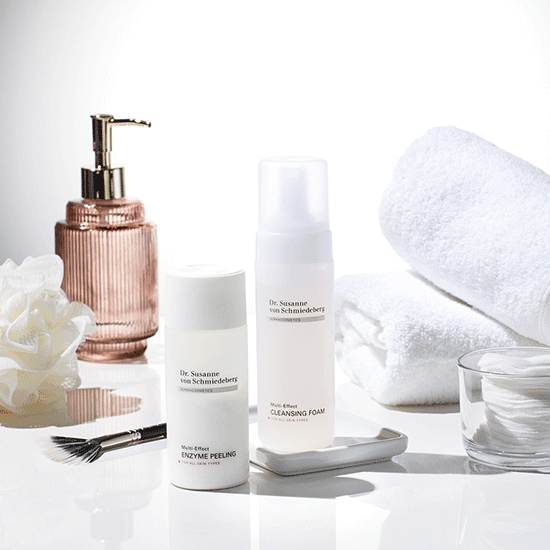 Se muestra un conjunto de productos Dr. Susanne von Schmiedeberg, incluyendo un peeling enzimático y una espuma limpiadora, junto con una toalla blanca y un dispensador de jabón dorado.
