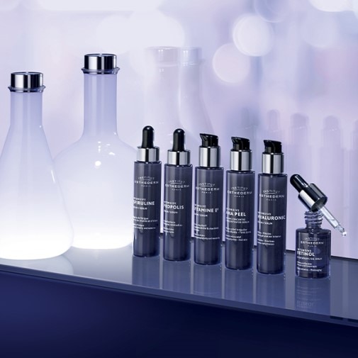 Kolekcja serum Institut Esthederm w ciemnych, eleganckich buteleczkach z pipetami, ustawiona na błyszczącej powierzchni, obok szklanych naczyń laboratoryjnych.