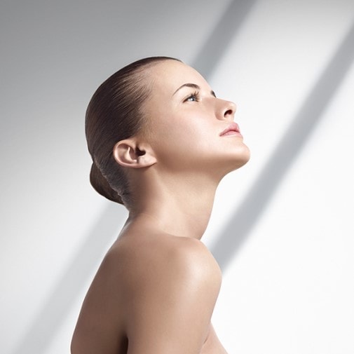 Portret kobiety z gładko zaczesanymi włosami, spoglądającej w górę na tle białej ściany z delikatnym cieniem.