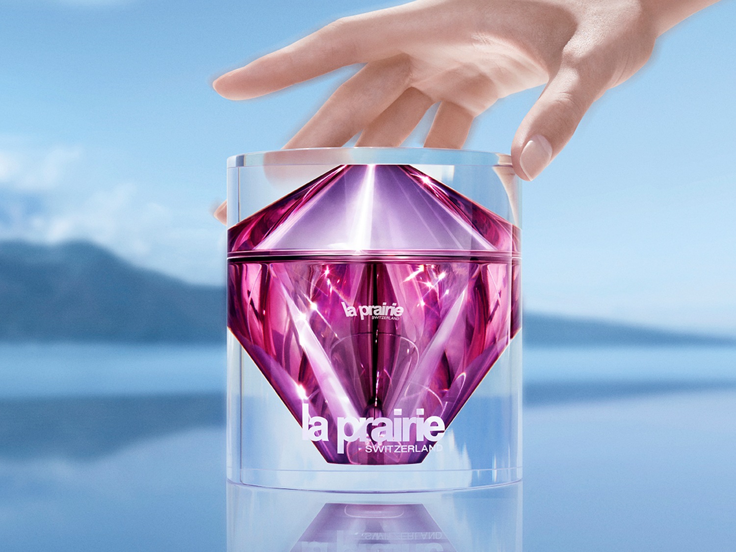 Detailní pohled na La Prairie pleťový krém v průhledném obalu s fialovým vnitřkem ve tvaru diamantu, ruka se ho dotýká, pozadí tvoří modrá obloha a jezero.
