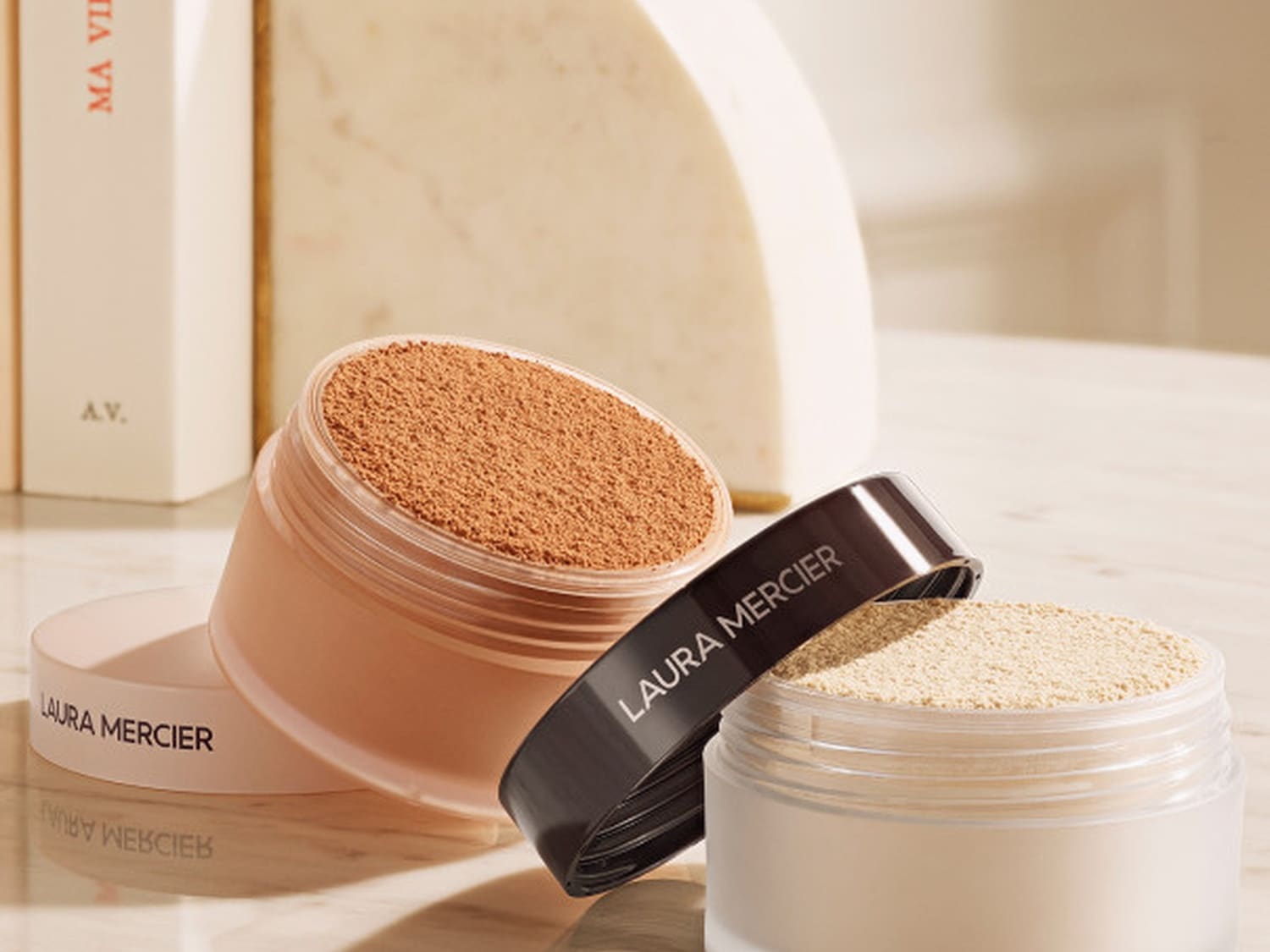 Dwa słoiczki pudru Laura Mercier: jeden z brązowym pudrem, drugi z beżowym, oba z otwartymi wieczkami, ukazujące sypką konsystencję.
