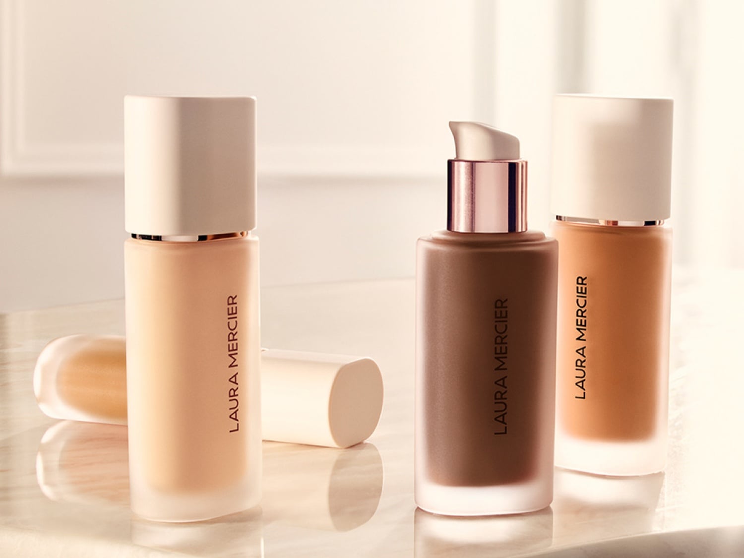 Trzy butelki podkładu Laura Mercier w różnych odcieniach, od jasnego beżu po głęboki brąz, z matowymi nakrętkami i eleganckim brandingiem.