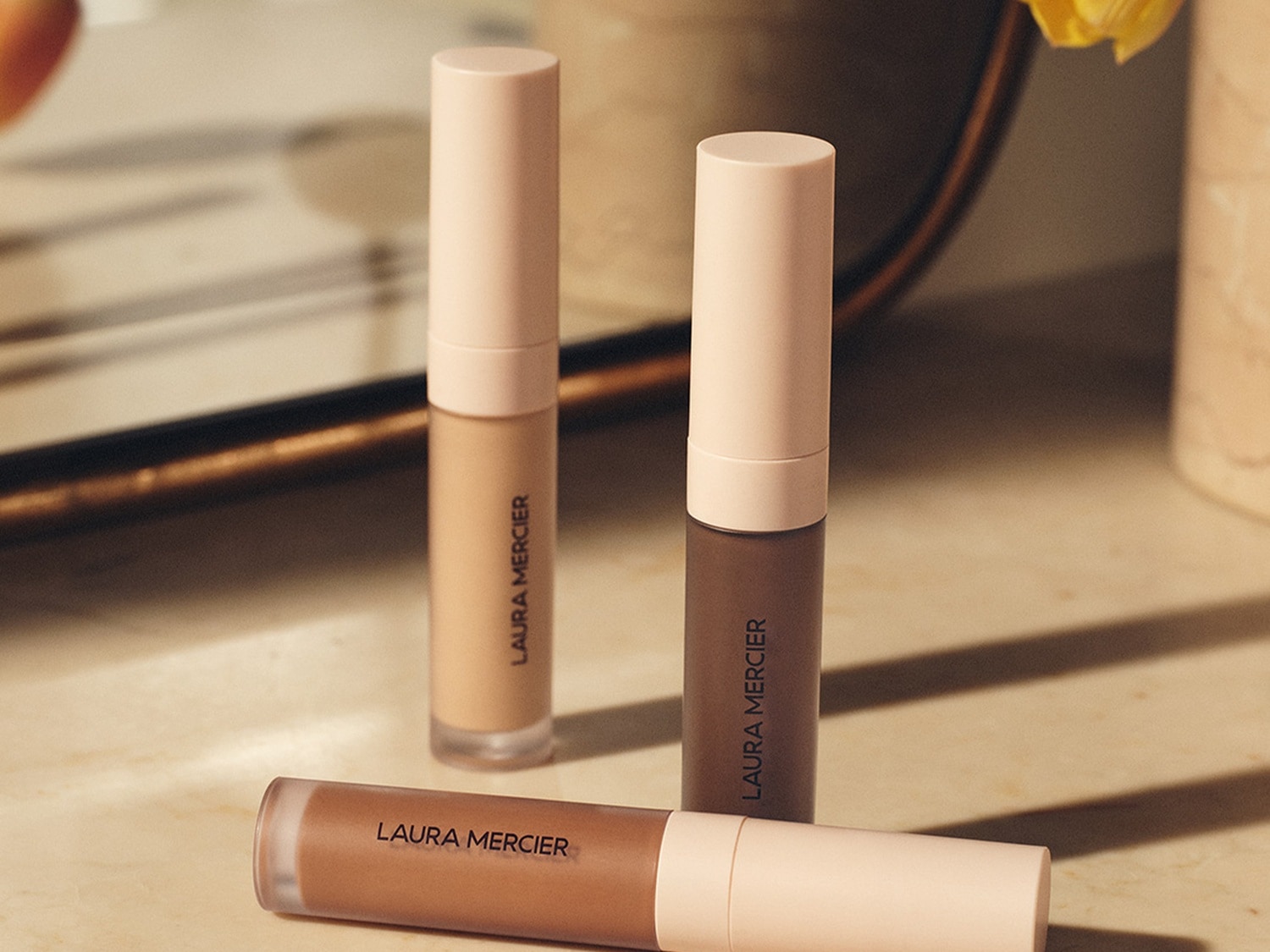 Trzy tubki korektora Laura Mercier w odcieniach beżu i brązu z jasnoróżowymi zakrętkami, leżące na blacie.