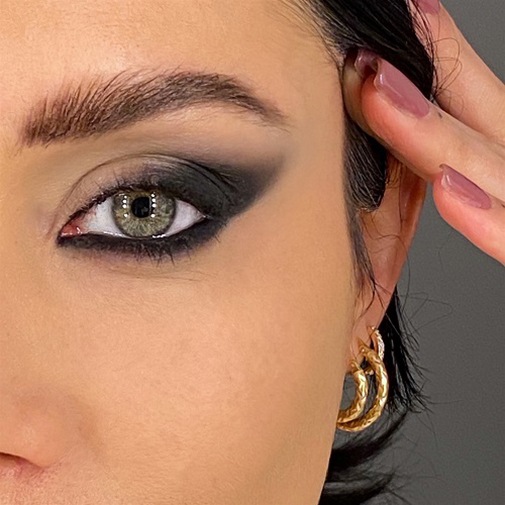 Primo piano di un occhio truccato con ombretto nero sfumato, sopracciglia definite e pelle chiara, completato da orecchini dorati.