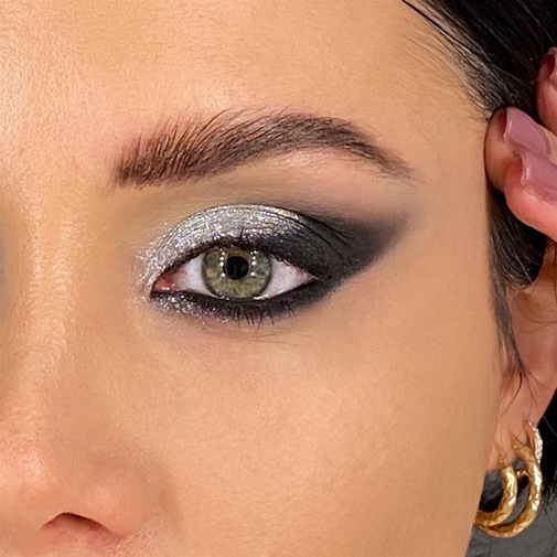 Primo piano di un occhio verde truccato con ombretto nero e argento glitterato, completato da un sopracciglio ben definito e un orecchino dorato.