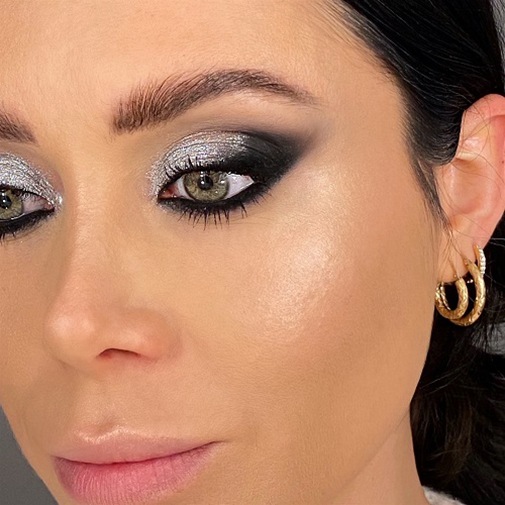 Primo piano di un volto femminile con un trucco occhi scintillante argento e nero, completato da orecchini dorati a cerchio.