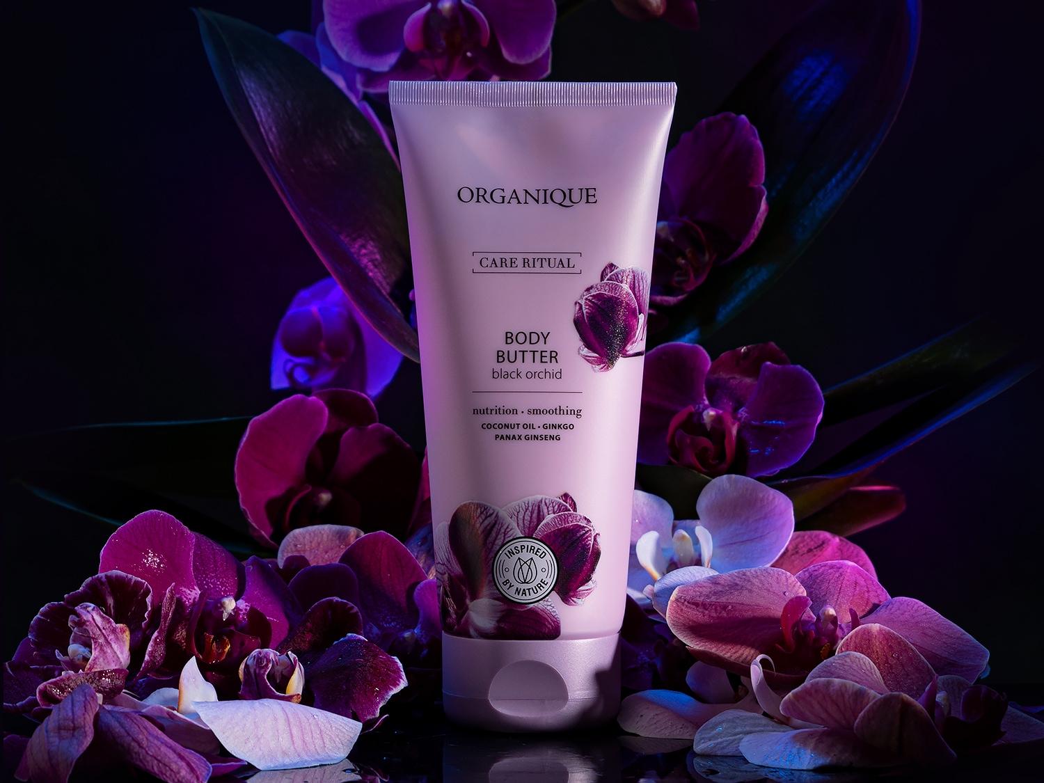 Fioletowa tubka masła do ciała Organique Care Ritual Black Orchid, otoczona kwiatami orchidei, obiecuje odżywienie i wygładzenie skóry dzięki zawartości oleju kokosowego, miłorzębu i żeń-szenia.