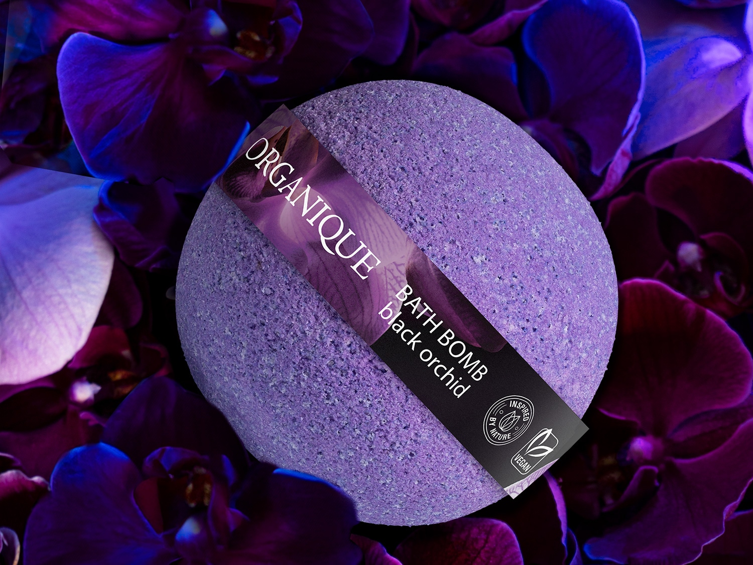 Fioletowa kula do kąpieli Organique Black Orchid otoczona kwiatami orchidei, etykieta informuje, że produkt jest wegański i inspirowany naturą.