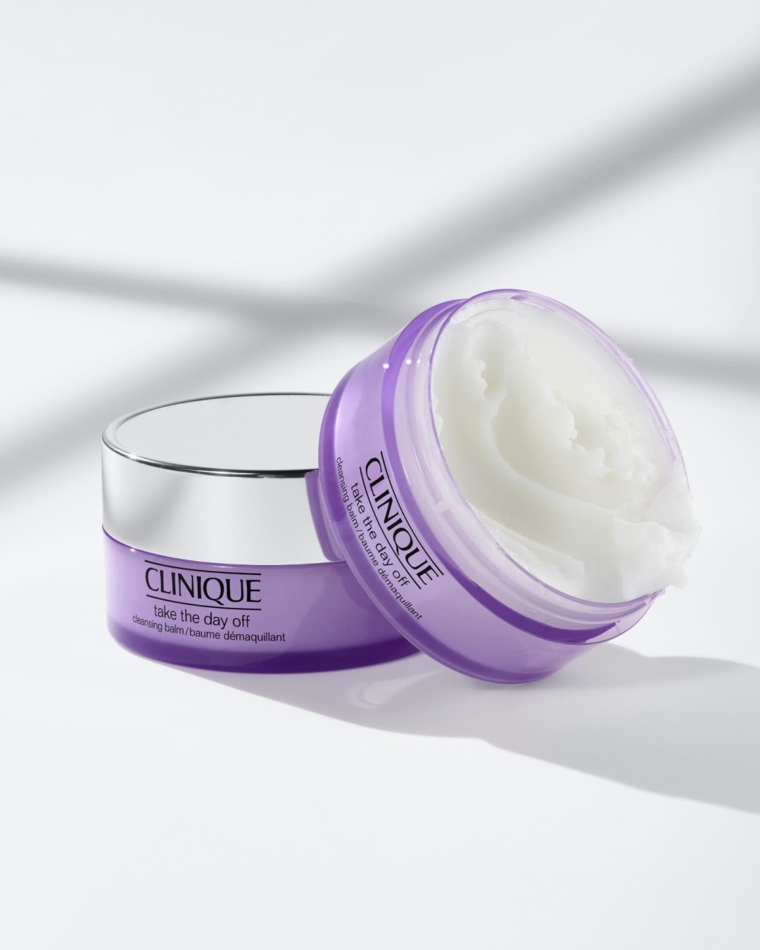 Dwa fioletowe słoiczki Clinique Take The Day Off: jeden zamknięty srebrnym wieczkiem, drugi otwarty, ukazujący biały, kremowy balsam do demakijażu.