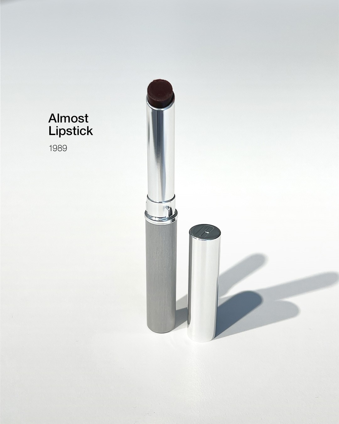 Szminka Clinique Almost Lipstick w srebrnym opakowaniu z brązowym odcieniem, obok leży srebrna nakrętka.